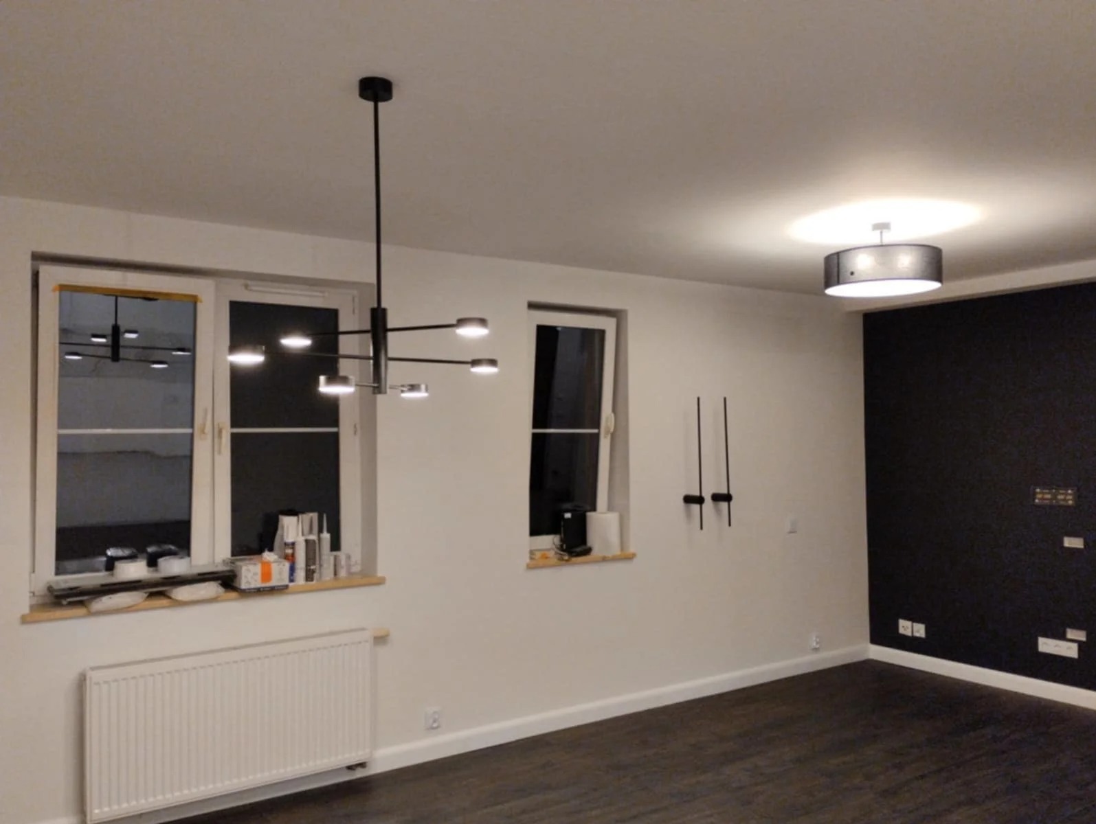 Nowoczesne wnętrze po remoncie: ciemna podłoga, białe ściany, czarna ściana akcentująca, okna z parapetami i designerskie lampy sufitowe.