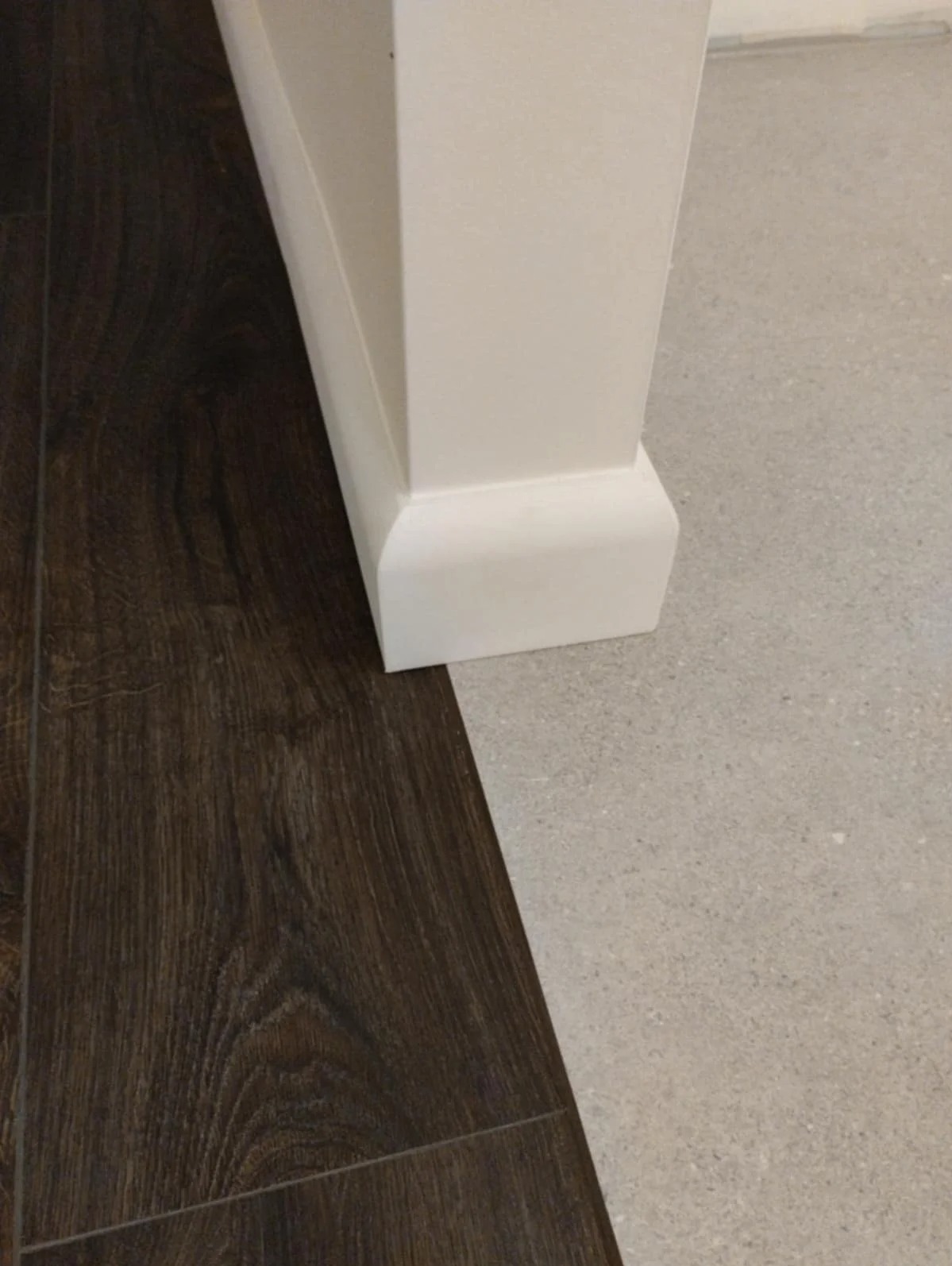 Połączenie dwóch rodzajów podłóg: paneli imitujących ciemne drewno i jasnych płytek. Biała listwa przypodłogowa. Ujęcie z bliska, ukazujące precyzję łączenia materiałów.