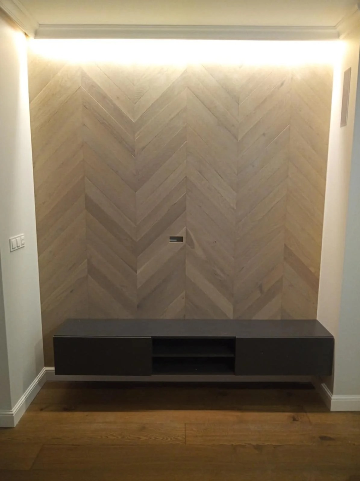 Ściana wykończona drewnianą jodełką z podświetleniem LED i zawieszona szafka RTV w nowoczesnym salonie. Parkiet na podłodze.