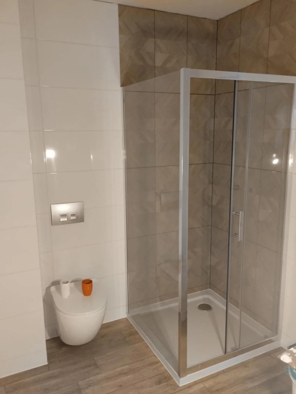 Nowoczesna kabina prysznicowa z przesuwnymi drzwiami, białe i beżowe płytki w łazience, drewnopodobna podłoga, wisząca toaleta z akcentem kolorystycznym.