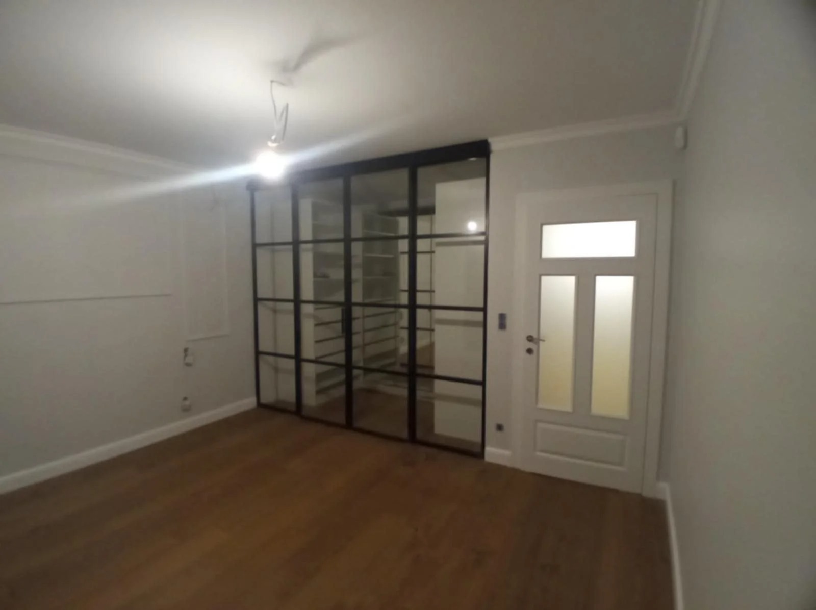 Nowoczesne wnętrze z drewnianą podłogą i przeszkloną ścianą działową z czarnymi szprosami, obok białe drzwi. Minimalistyczny design, jasne ściany.