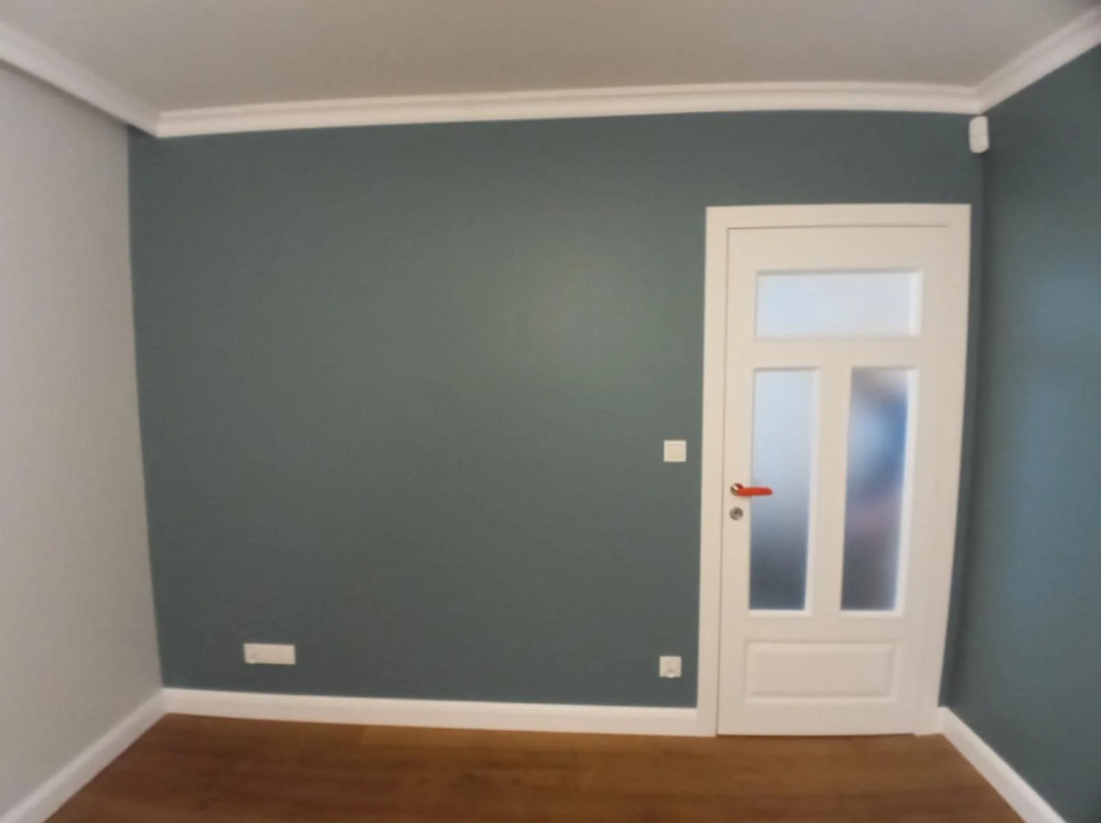 Pomieszczenie po malowaniu ścian na kolor morski z białą stolarką drzwiową i przypodłogową. Widoczna drewniana podłoga i biały sufit z dekoracyjną listwą.