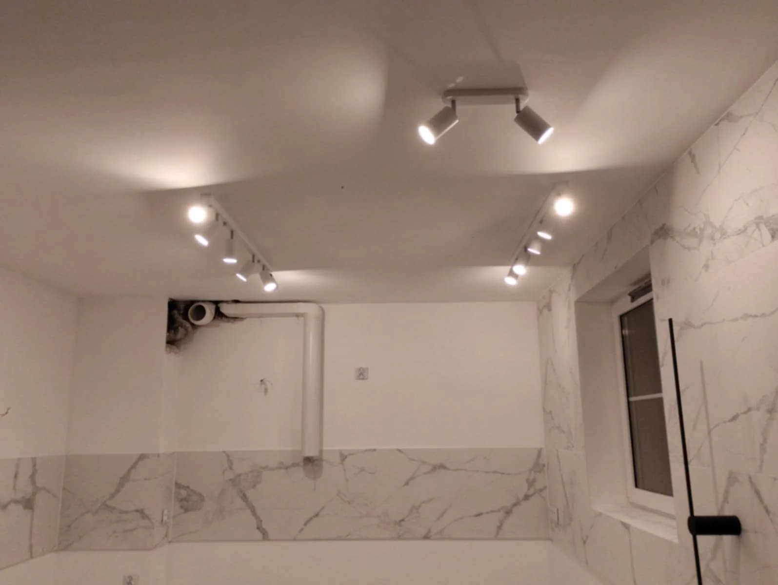 Wykończona łazienka z marmurowymi płytkami i nowoczesnym oświetleniem punktowym. Widoczne detale instalacji pod sufitem. Minimalistyczny design.