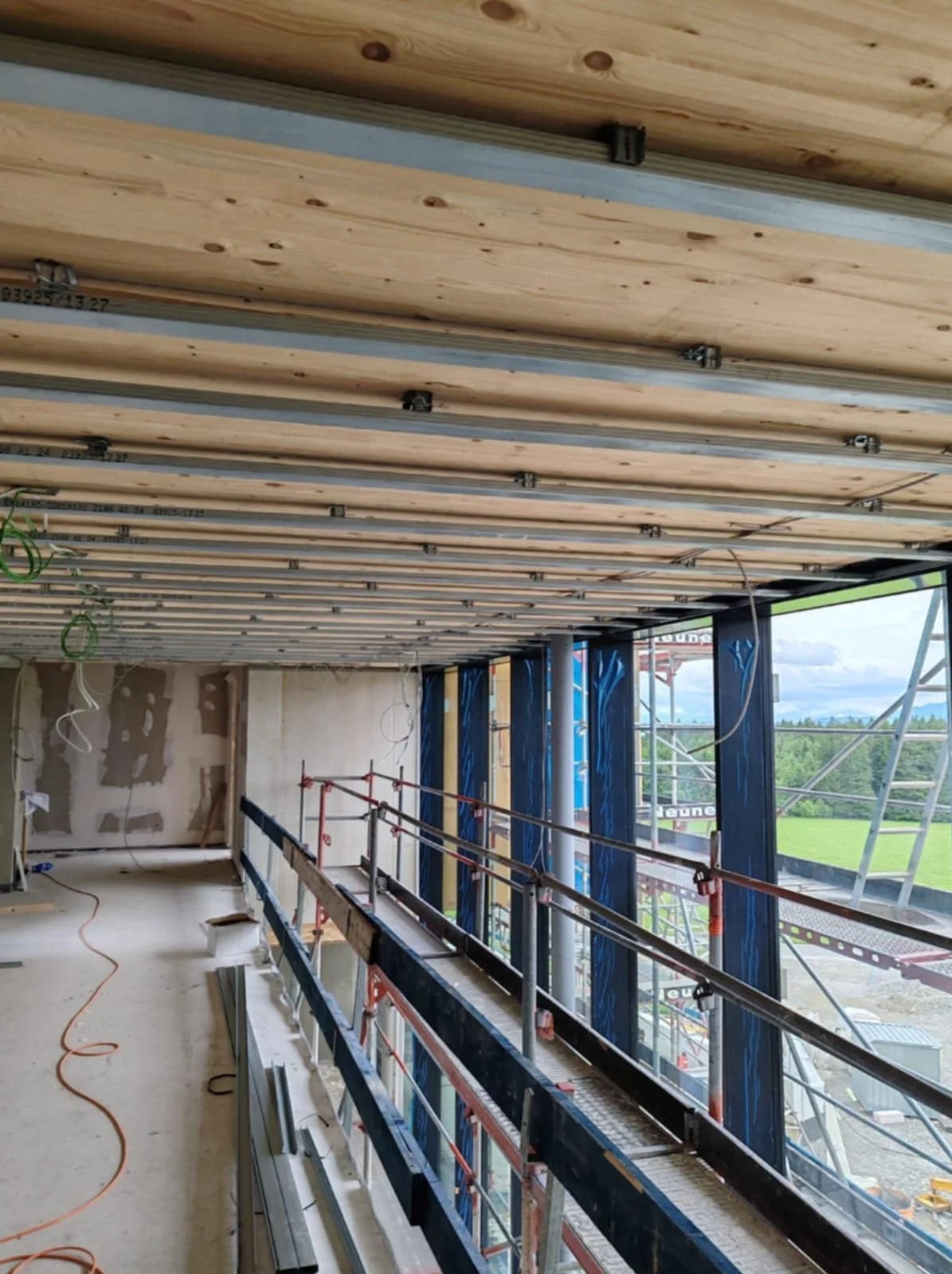 Wnętrze w trakcie wykończenia: drewniany sufit z metalowymi profilami, ściany w stanie surowym, rusztowania, duże okna z widokiem na zieleń. Prace wykończeniowe w nowoczesnym budynku.