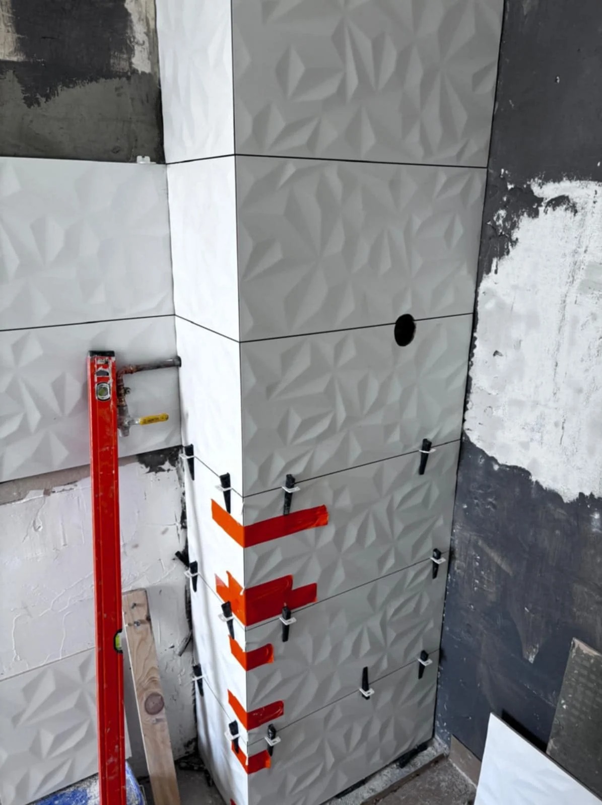 Białe, trójwymiarowe płytki ułożone na ścianie, z użyciem poziomicy i klipsów dystansowych. Widoczne surowe wykończenie ściany obok.