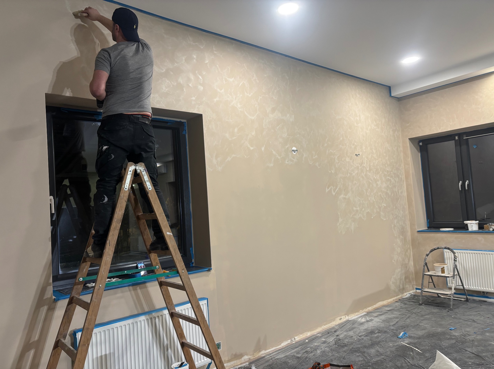 Mężczyzna na drabinie maluje ścianę z efektem dekoracyjnym w beżowych odcieniach. Okna zabezpieczone taśmą malarską. Widoczne narzędzia malarskie i folia ochronna na podłodze.