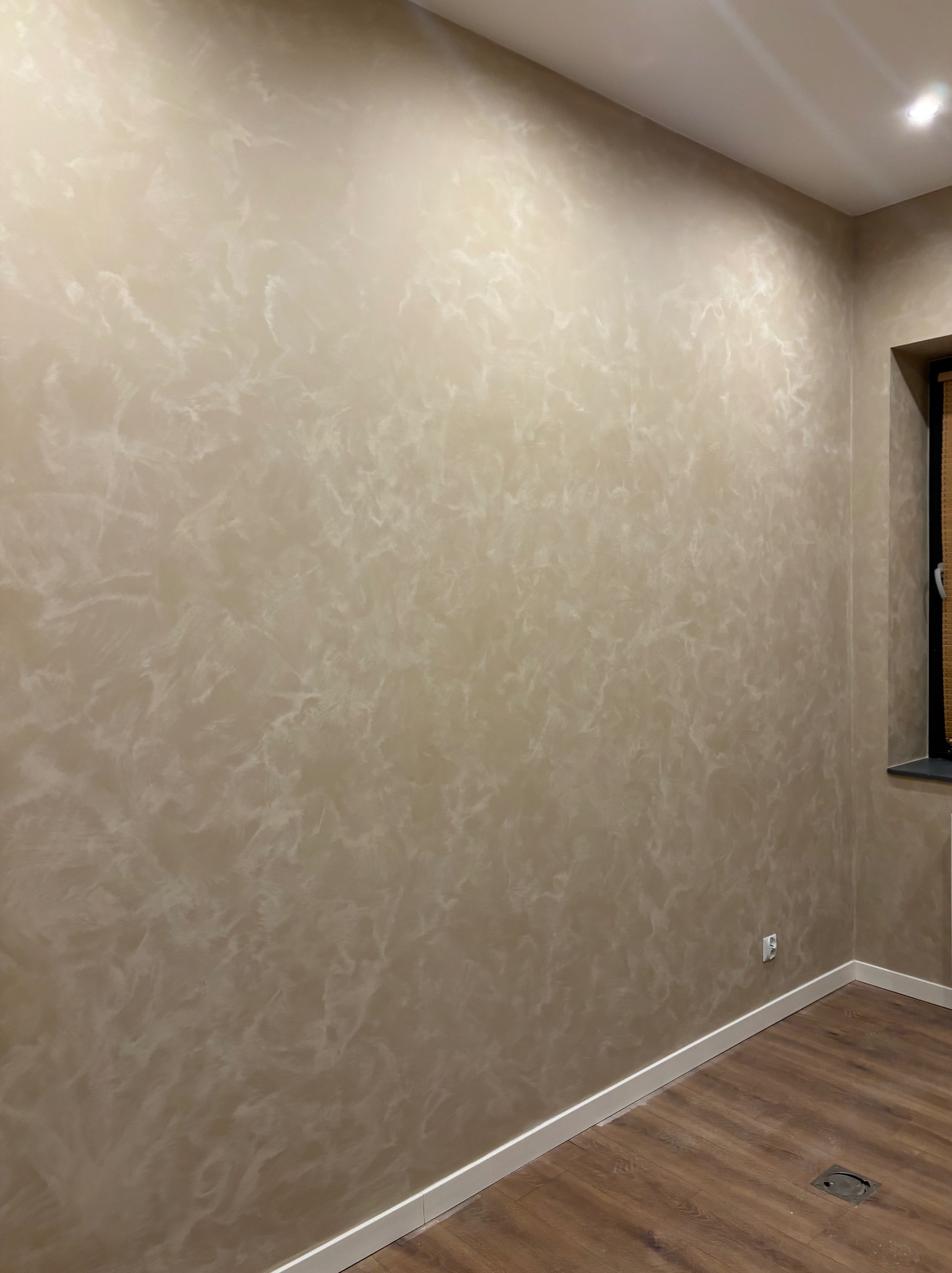 Beżowa ściana z efektem dekoracyjnym w wykończonym pokoju, widoczna listwa przypodłogowa i fragment drewnianej podłogi. Ujęcie z bliska prezentuje fakturę ściany.