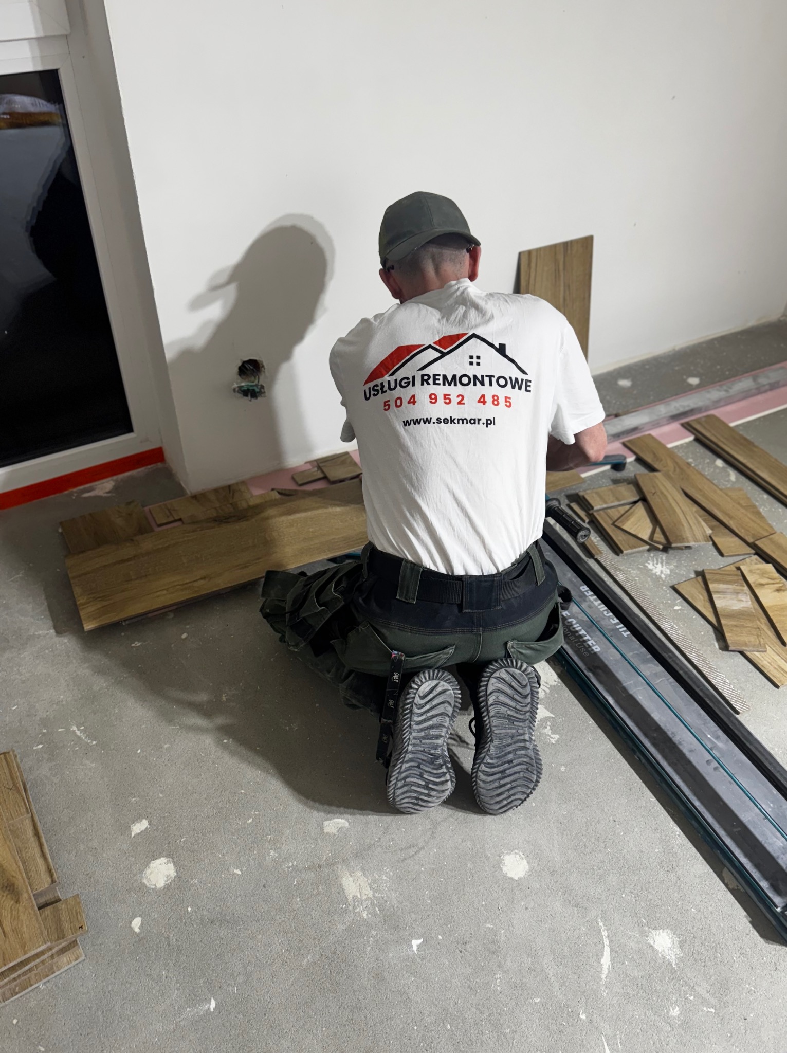 Pracownik w firmowej koszulce układa panele podłogowe w nowym pomieszczeniu. Widoczny profesjonalny sprzęt do cięcia paneli i przygotowane deski.