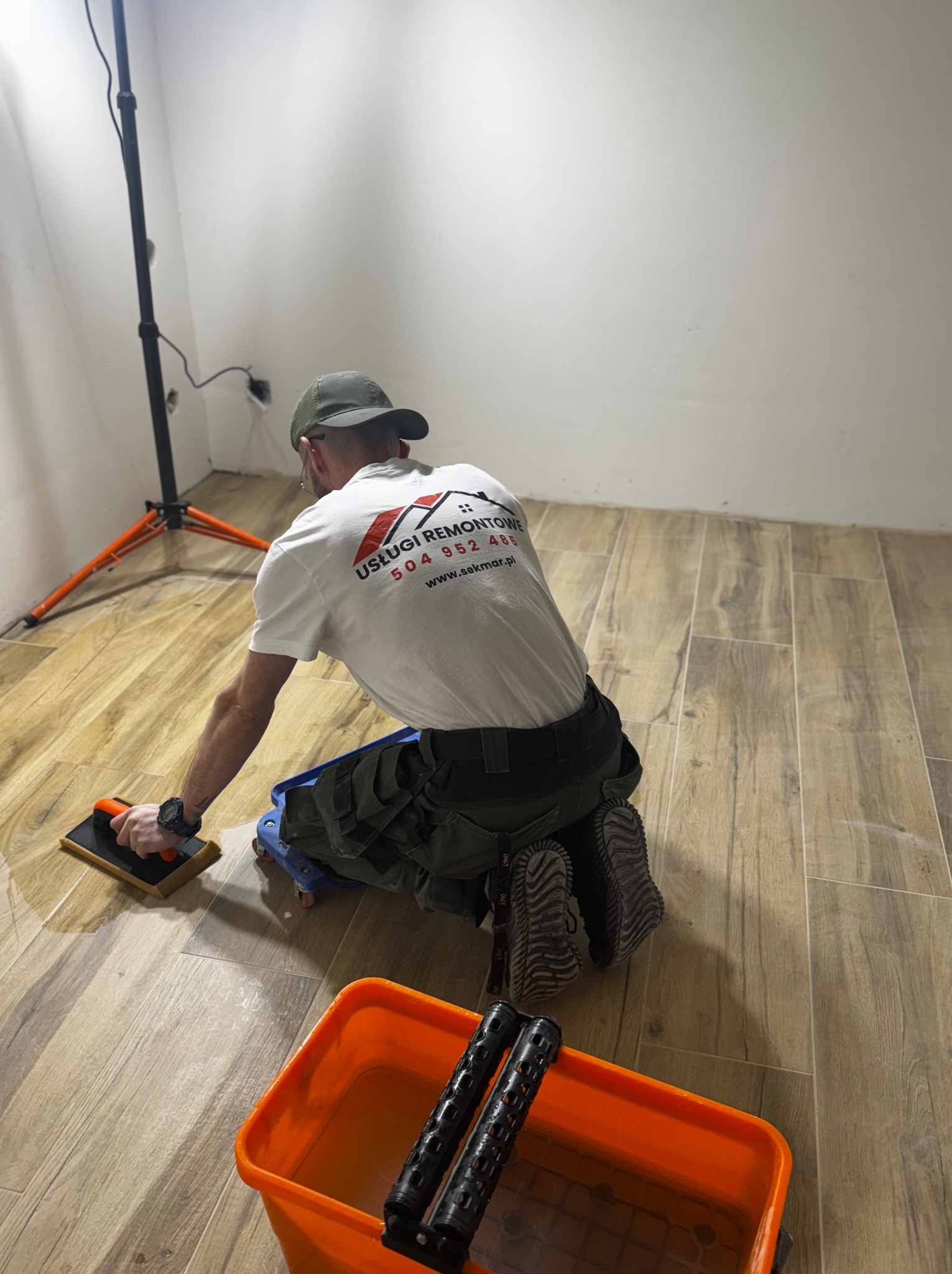 Glazurnik na kolanach czyści świeżo ułożone, podłużne płytki podłogowe imitujące drewno. Widoczny sprzęt do czyszczenia i wiadro. Na koszulce logo firmy.