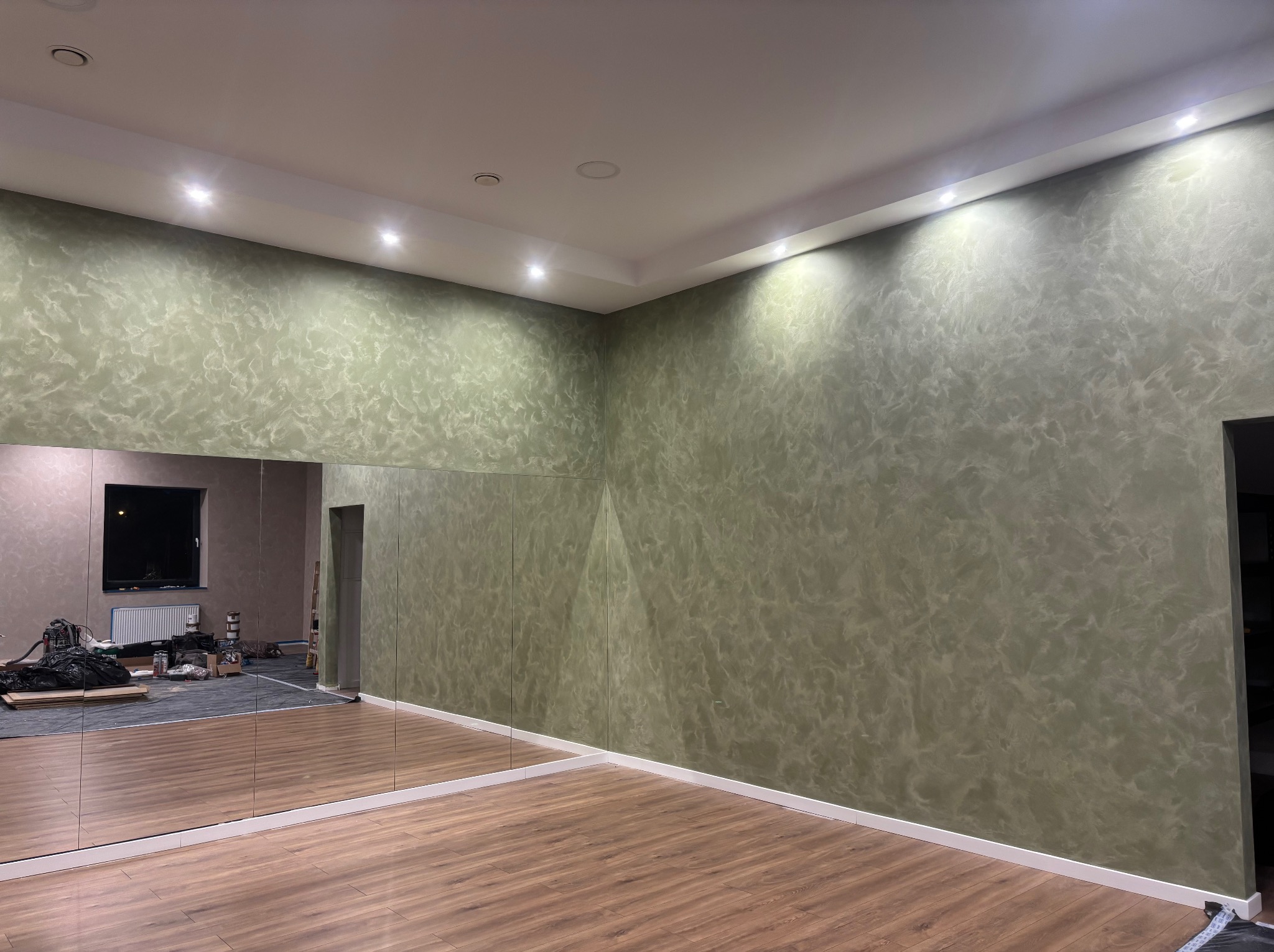 Wykończone wnętrze: ściana z dekoracyjnym malowaniem w odcieniach zieleni, lustra na ścianie, drewniana podłoga i oświetlenie punktowe. Widok na salę taneczną.