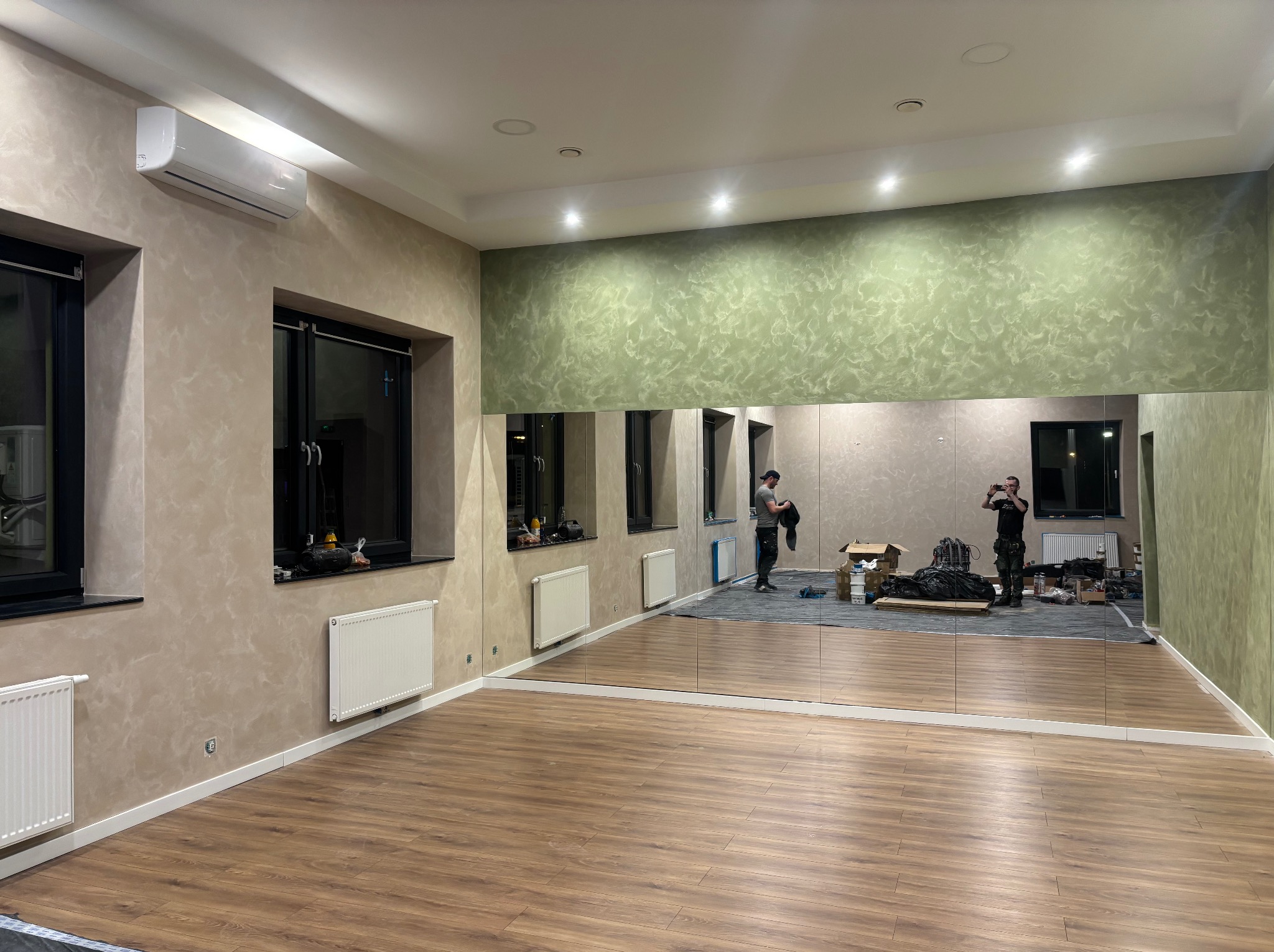 Sala po odświeżeniu: drewnopodobna podłoga, lustra na ścianie, beżowe i zielone ściany z efektem dekoracyjnym, klimatyzator. Widoczni pracownicy podczas prac wykończeniowych.