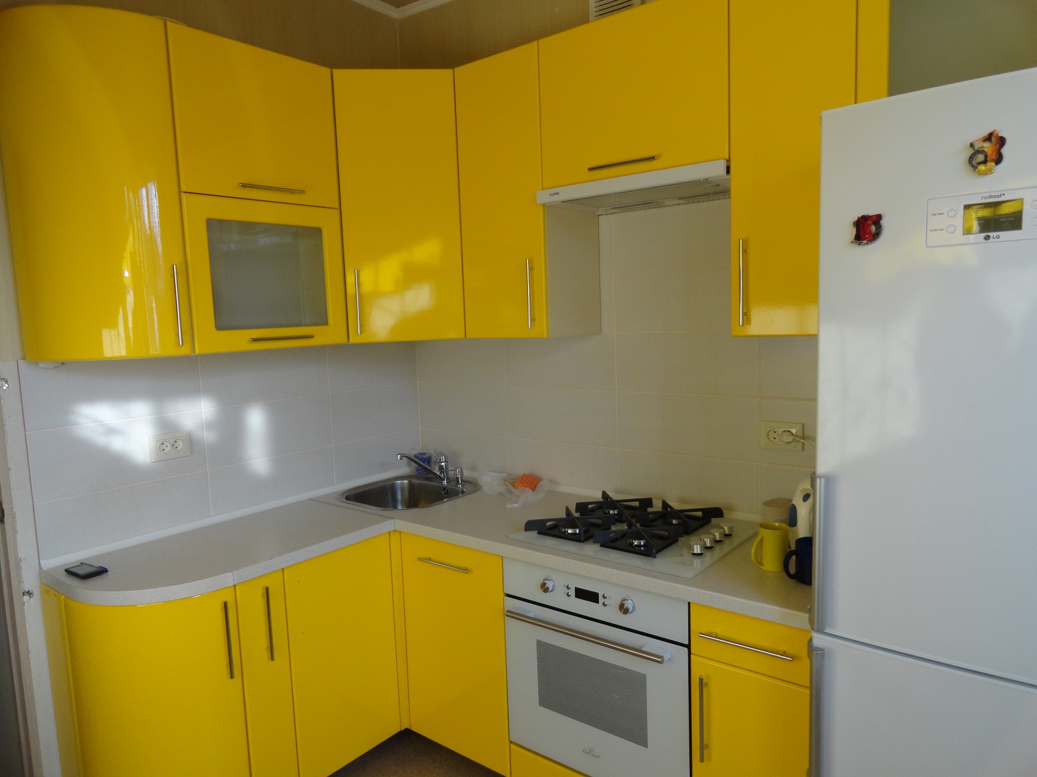 Żółta kuchnia z białymi blatami i sprzętem AGD, w tym płytą grzewczą i lodówką. Uwagę przykuwają magnetyczne ozdoby na lodówce.