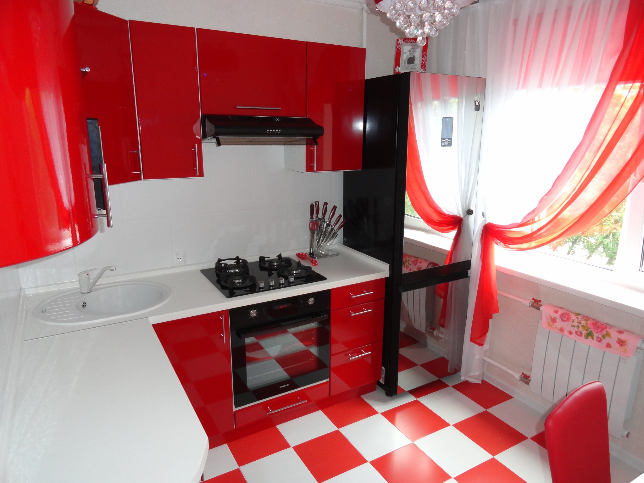 Czerwono-biała kuchnia z błyszczącymi frontami, czarnym sprzętem AGD i podświetleniem. Uwagę przykuwają okiennice i wzorzysta podłoga.