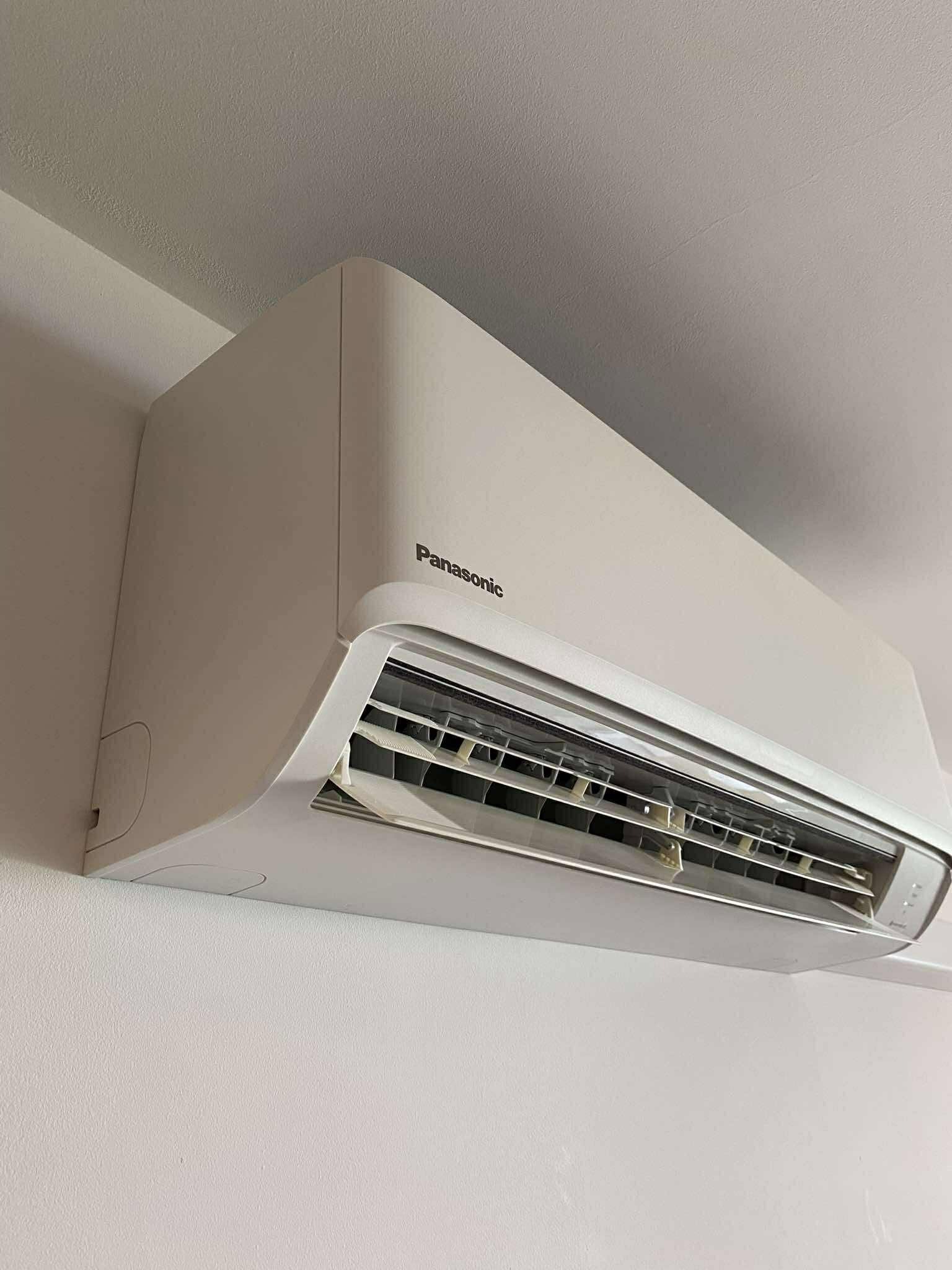 Zamontowana, biała klimatyzacja Panasonic na jasnej ścianie, widok z dołu. Widoczne żaluzje wewnątrz urządzenia i fragment sufitu.