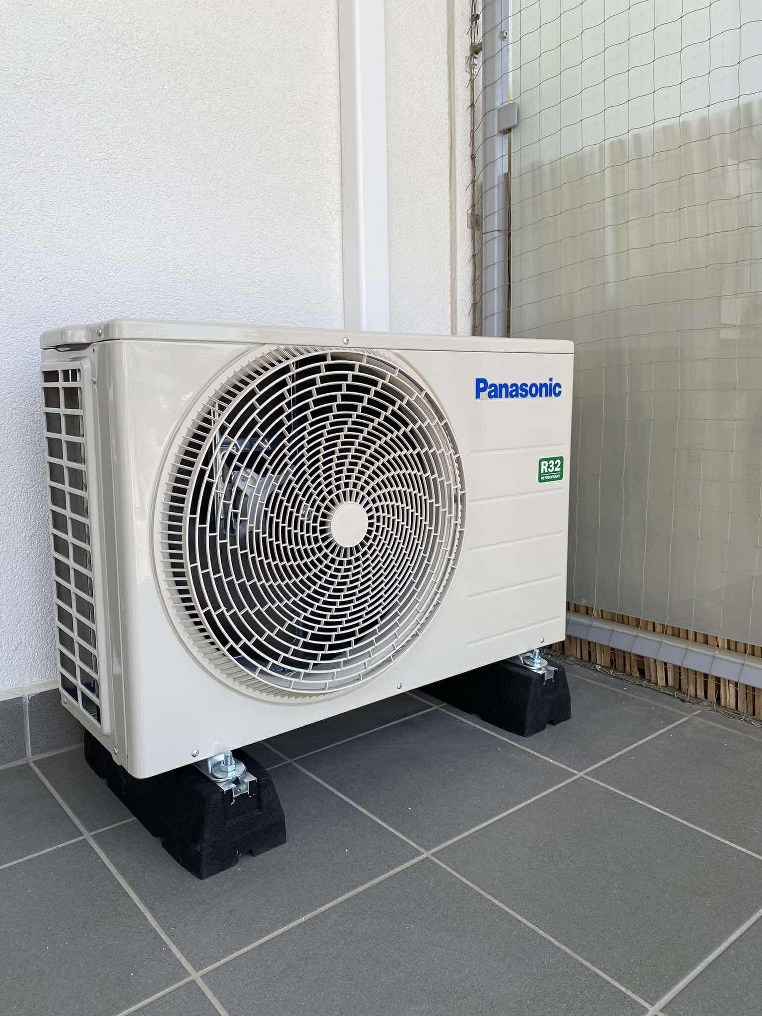 Zewnętrzna jednostka klimatyzacji Panasonic na czarnych podkładkach antywibracyjnych, ustawiona na szarych płytkach tarasu przy białej ścianie budynku.