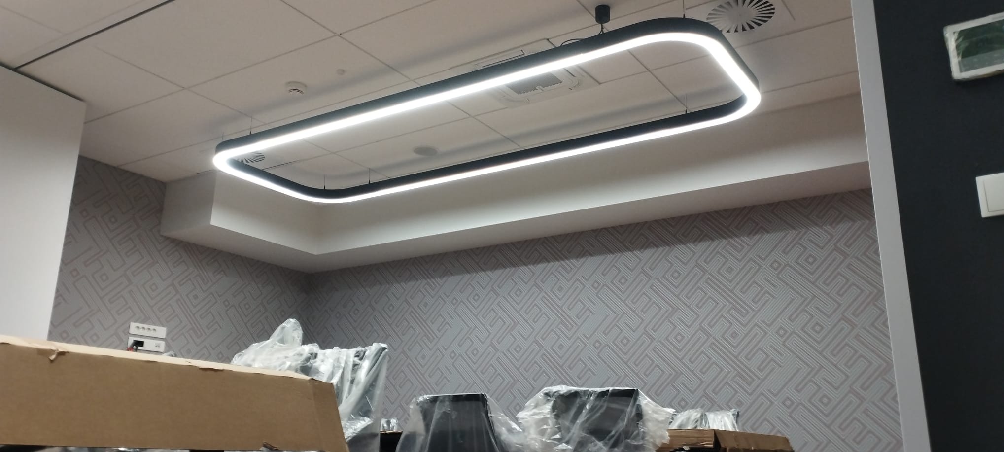 Nowoczesna lampa sufitowa LED w kształcie prostokąta z zaokrąglonymi rogami, zawieszona w biurze z wzorzystą tapetą. Widoczne kartony i krzesła w folii.