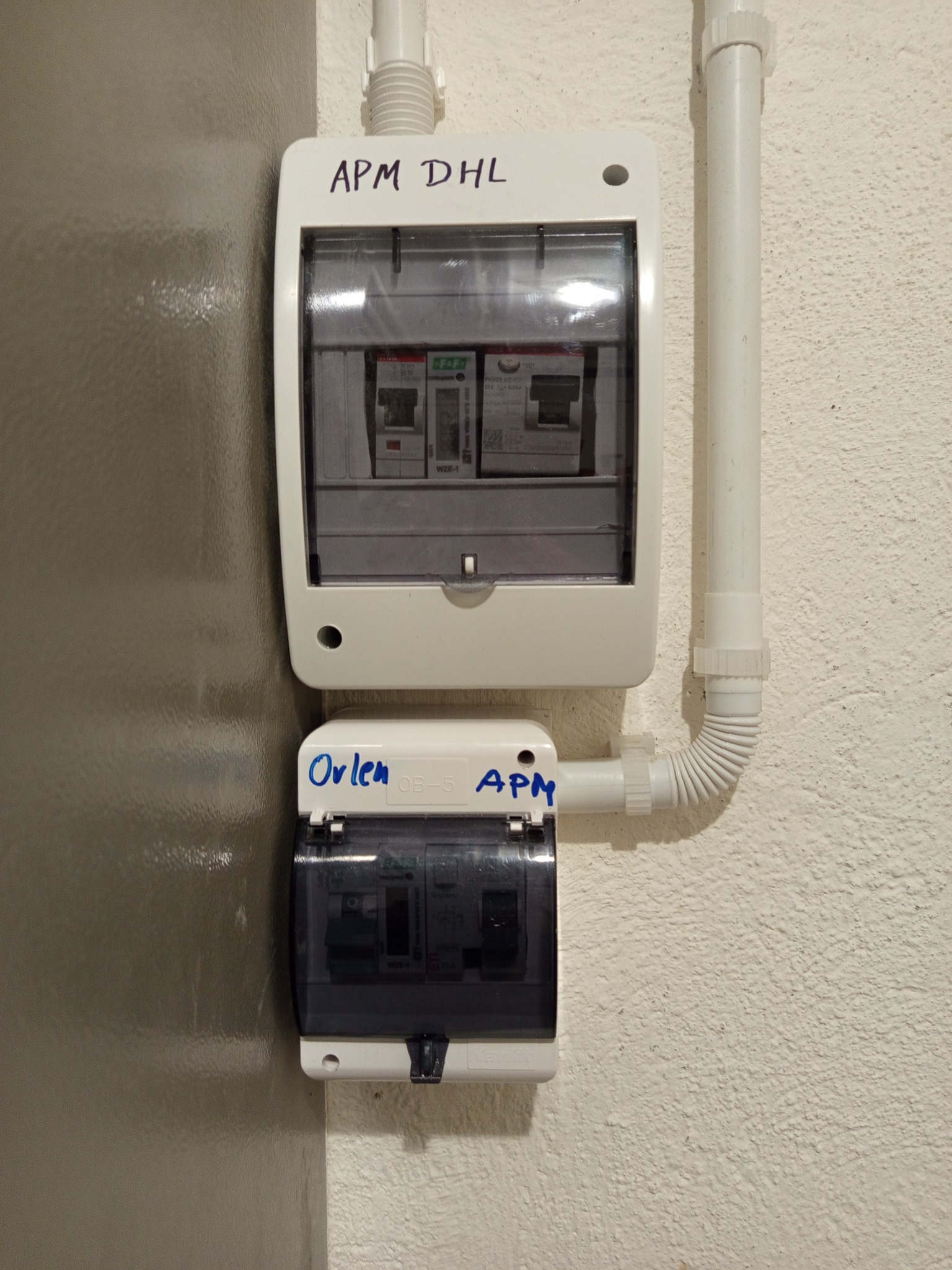 Dwa białe rozdzielacze elektryczne z bezpiecznikami, zamontowane na ścianie obok rury. Na obudowach widoczne napisy wykonane markerem.