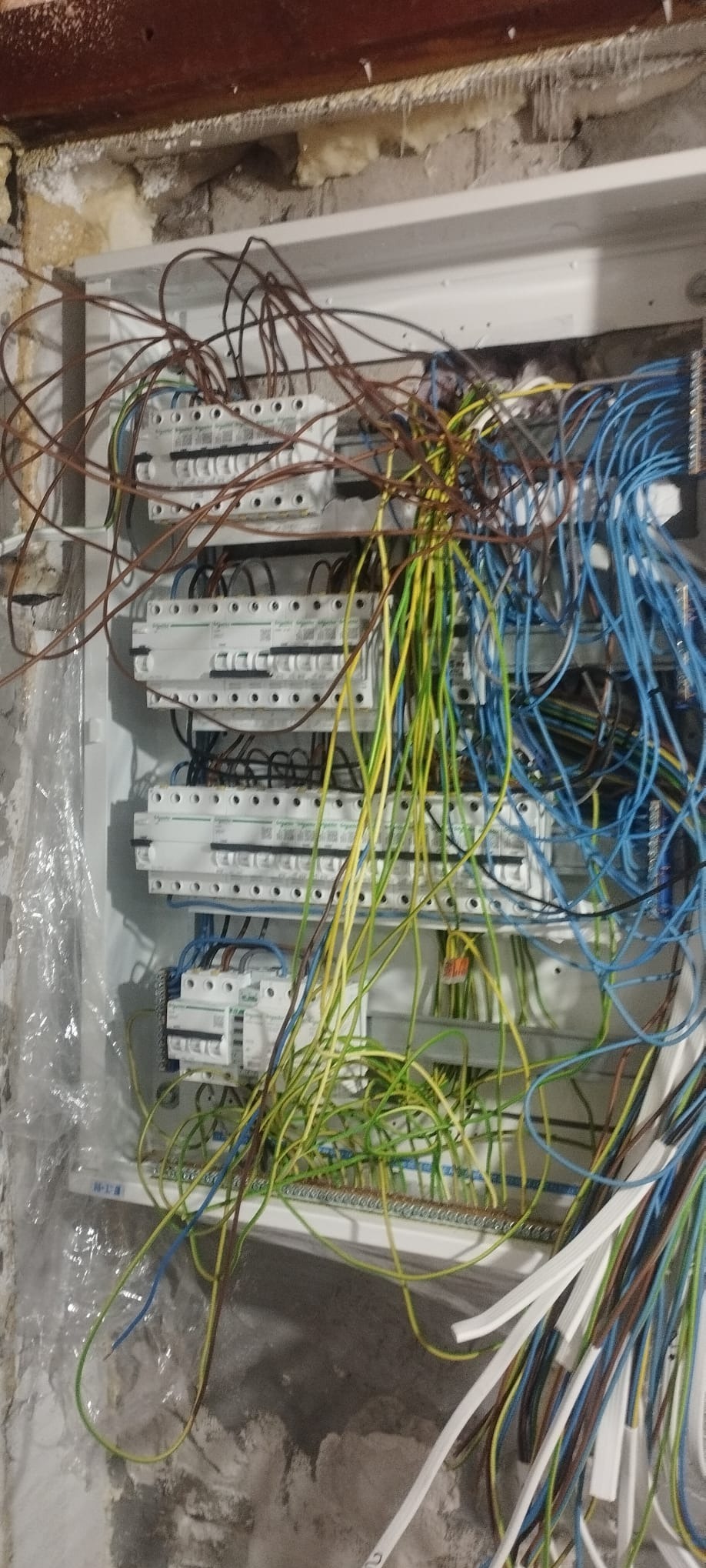 Rozdzielnia elektryczna w trakcie montażu, widoczne liczne przewody w różnych kolorach, biała obudowa i surowa ściana w tle. Nieuporządkowane okablowanie.