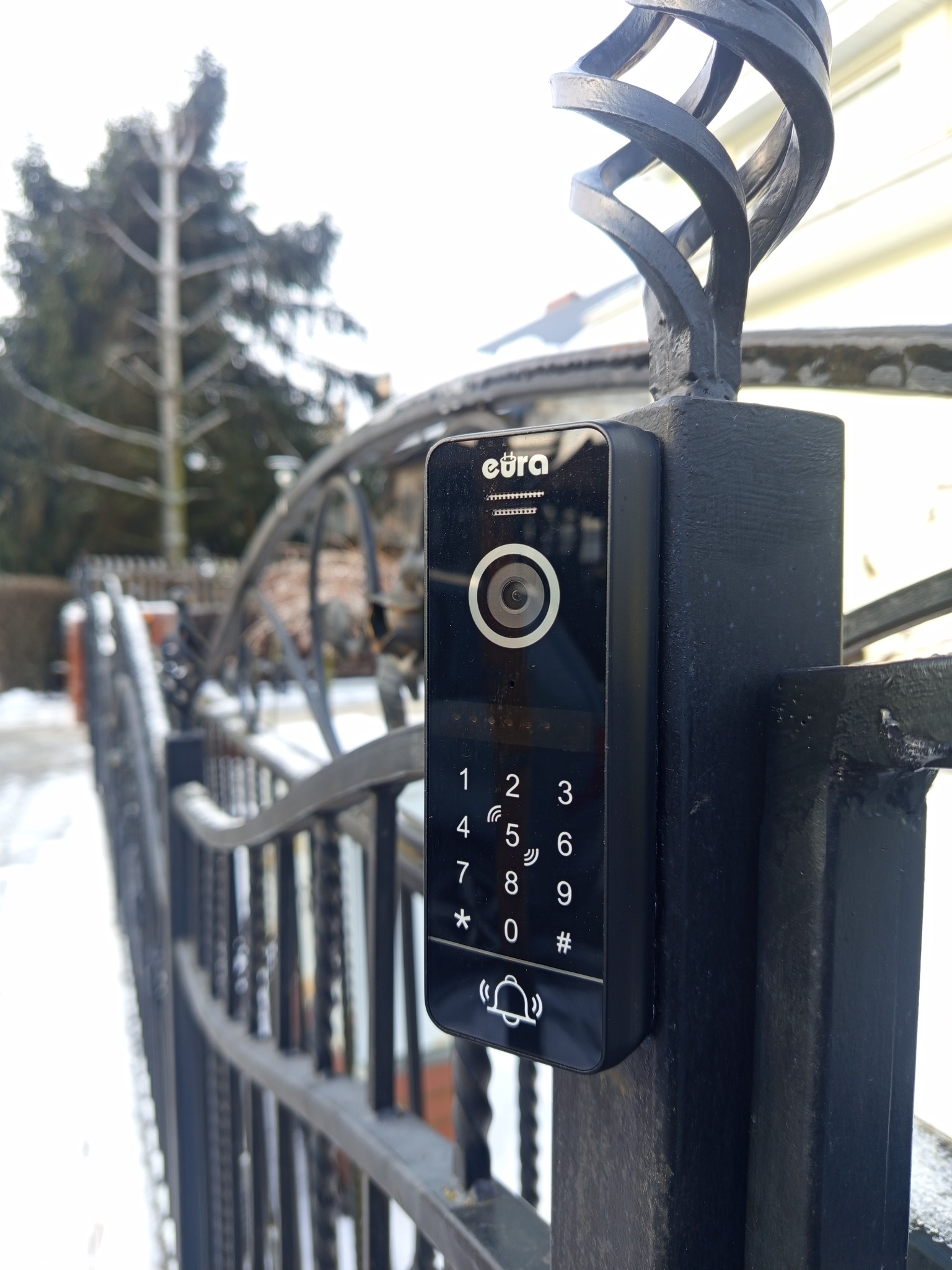 Zamontowany wideodomofon marki Eura na czarnym, metalowym słupku bramy, z widoczną klawiaturą numeryczną i kamerą. W tle brama i zimowy krajobraz.