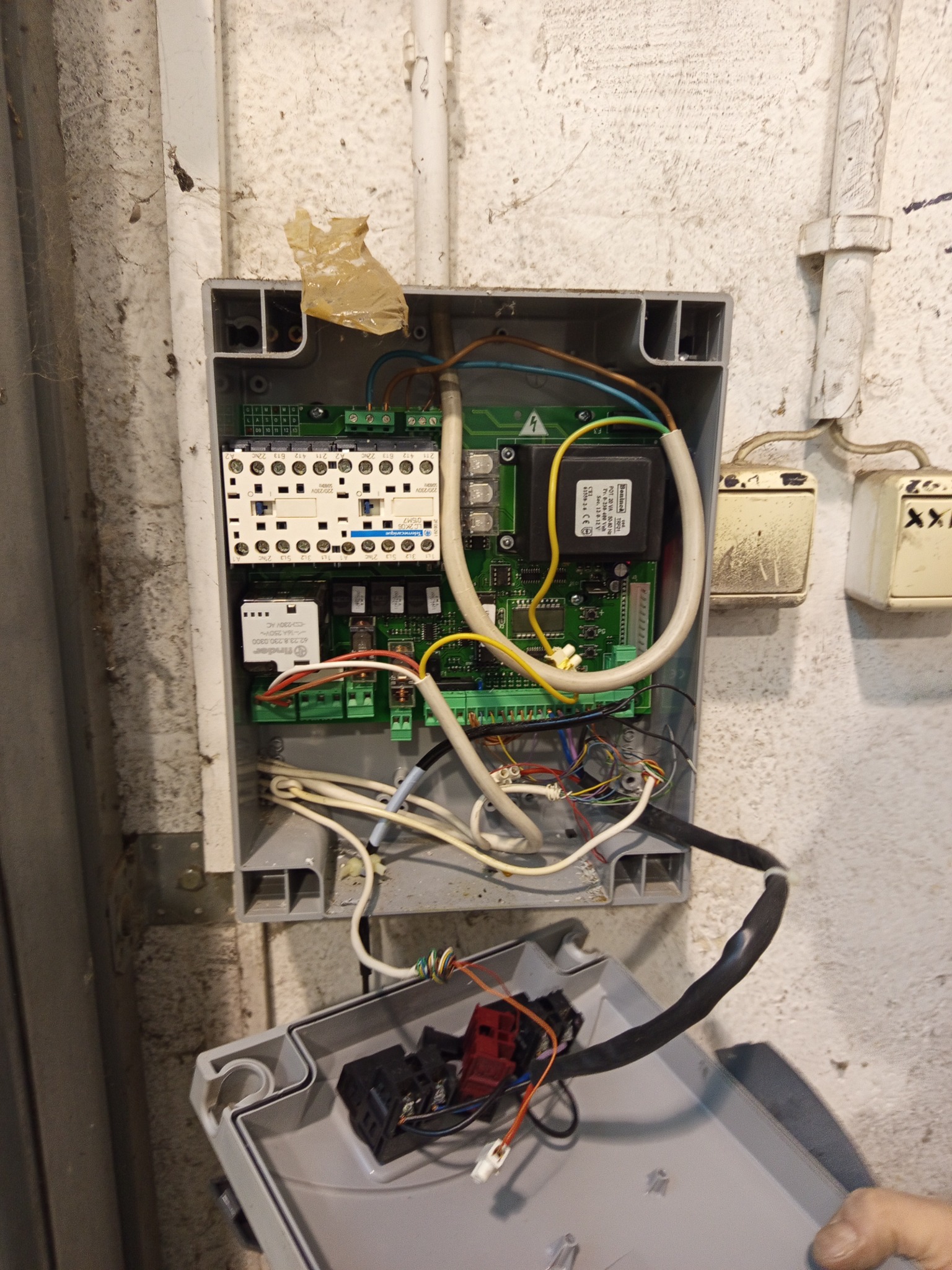 Otwarta skrzynka sterownicza automatyki bramy z widocznymi stycznikami, motoreduktorem i przewodami na tle ściany. Widoczne elementy elektryczne i elektronika.