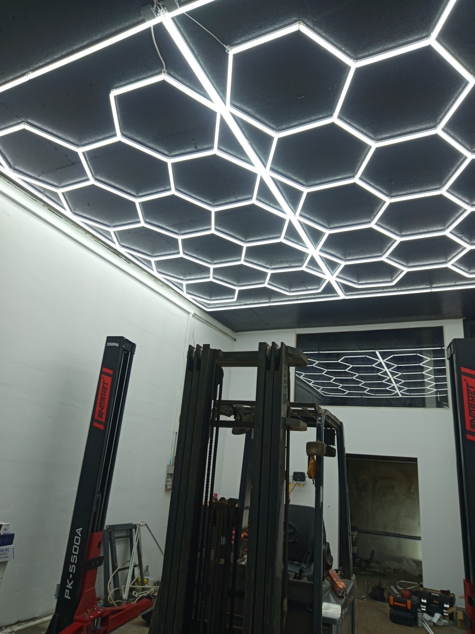 Warsztat z oświetleniem LED w kształcie heksagonów na suficie, widoczny podnośnik samochodowy i wózek widłowy. Jasne, równomierne światło.