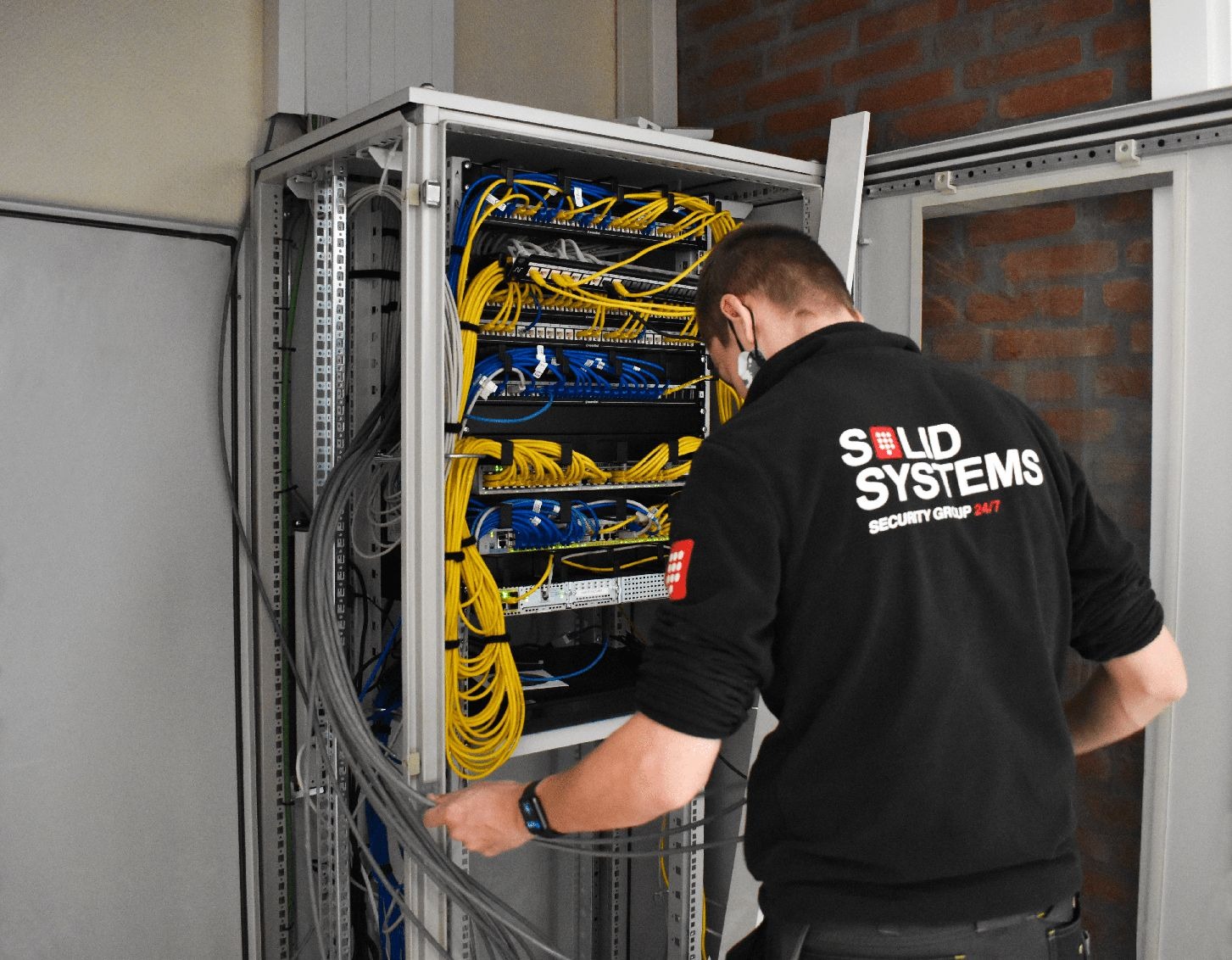 Instalator w czarnej bluzie z logo firmy układa kable w szafie rack z widocznymi patch panele i switchami. Ściana z czerwonej cegły w tle.