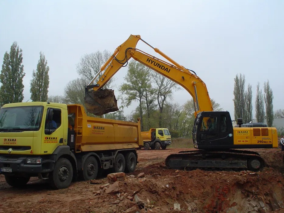 Żółty koparka Hyundai nabiera ziemię do wywrotki KAMA na tle wykopu i innych pojazdów. Praca ziemna w toku, widok z boku, pochmurny dzień.
