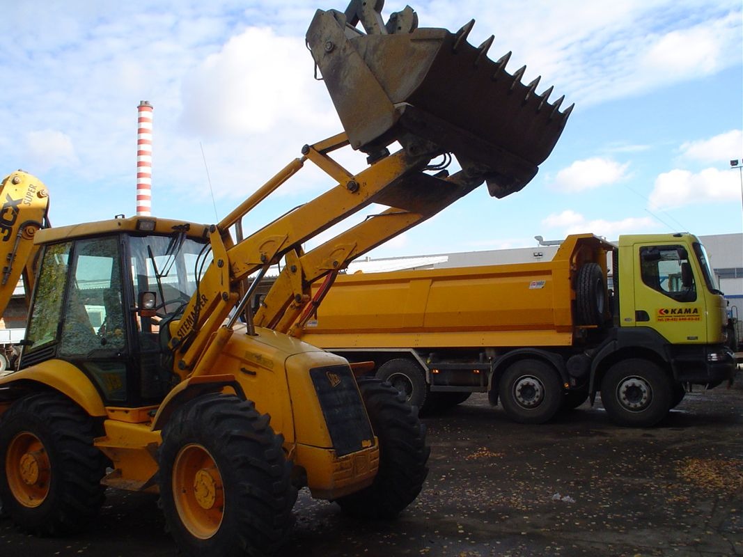 Żółta koparko-ładowarka JCB z podniesioną łyżką i wywrotka KAMA w tle, na placu budowy. W tle komin w biało-czerwone pasy.