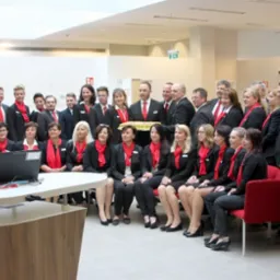 Grupowe zdjęcie pracowników banku Austria z logo na fladze i laptopie, ubranych w czarne uniformy z czerwonymi apaszkami, w jasnym, nowoczesnym wnętrzu.