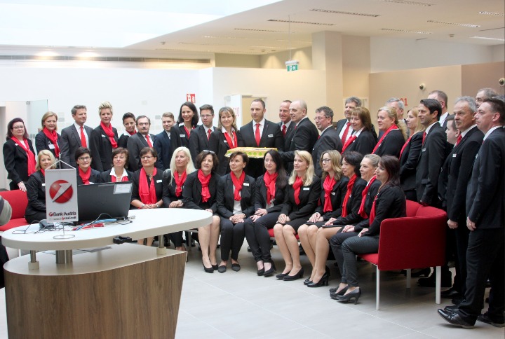 Grupowe zdjęcie pracowników banku Austria z logo na fladze i laptopie, ubranych w czarne uniformy z czerwonymi apaszkami, w jasnym, nowoczesnym wnętrzu.