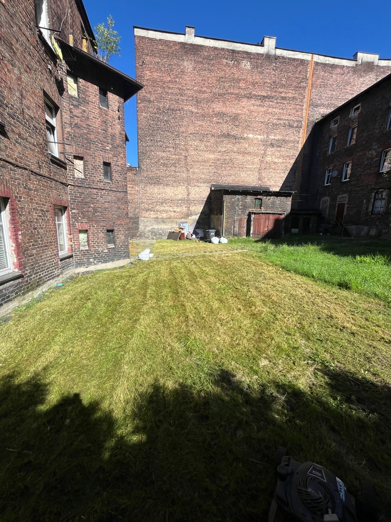 Świeżo skoszony trawnik z widocznymi pasami, otoczony ceglanymi budynkami w stylu industrialnym. W tle narzędzia ogrodnicze i sprzęt. W dolnej części kadru fragment kosiarki.