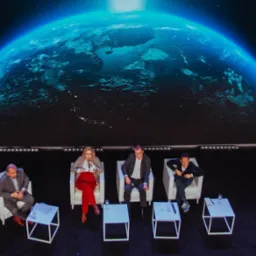Panel dyskusyjny w Warszawie. Cztery osoby siedzą na białych krzesłach na scenie z ekranem LED wyświetlającym grafikę Ziemi.