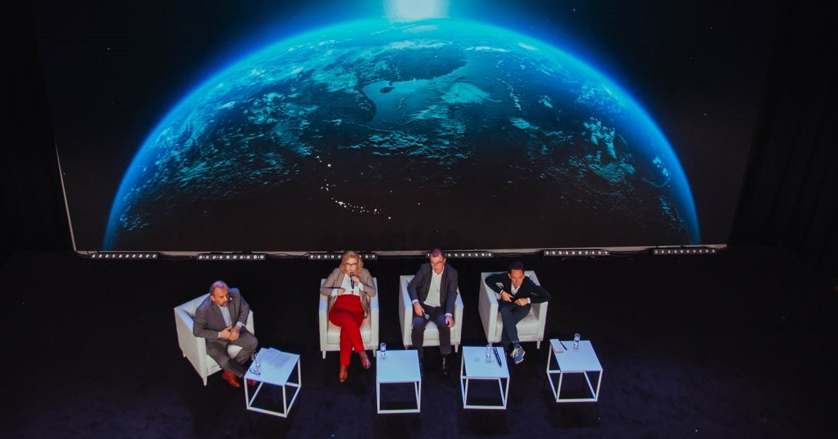 Panel dyskusyjny w Warszawie. Cztery osoby siedzą na białych krzesłach na scenie z ekranem LED wyświetlającym grafikę Ziemi.