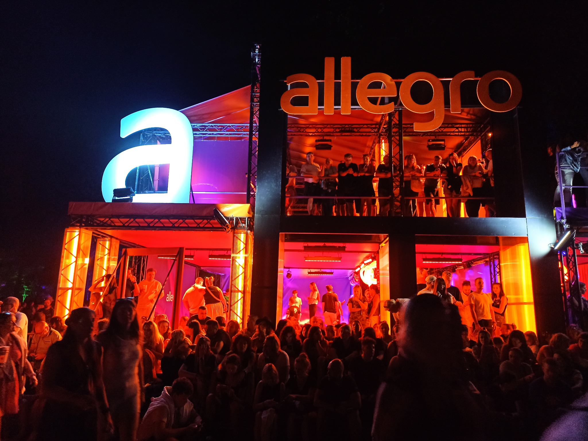 Scena z logotypem 'allegro', oświetlona na czerwono i fioletowo, z tłumem ludzi podczas wydarzenia. Widoczne osoby na różnych poziomach konstrukcji scenicznej.