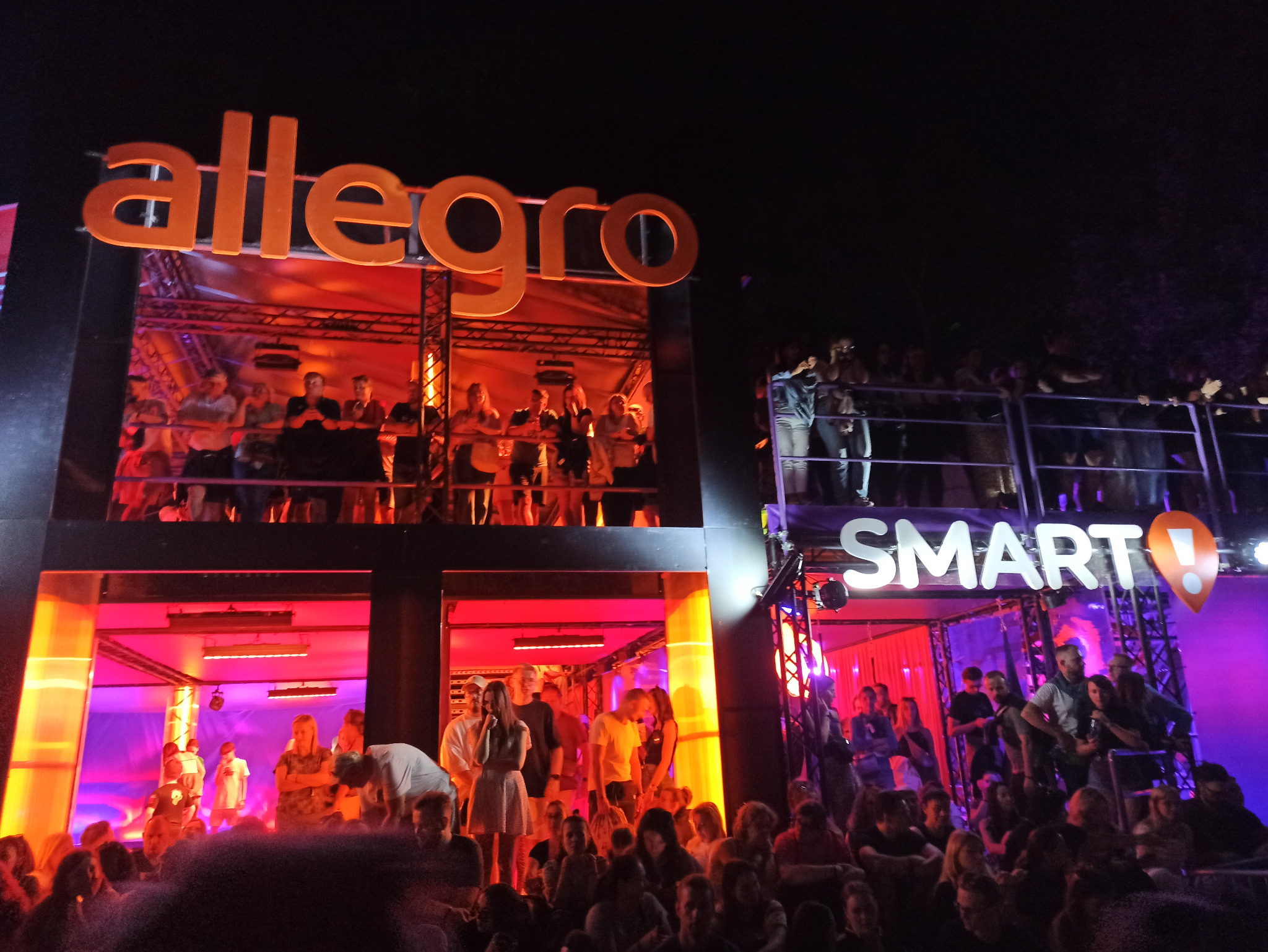 Event firmowy z logo Allegro i Smart, scena z oświetleniem, tłum ludzi. Wydarzenie wieczorne, dynamiczne ujęcie z dołu, ukazujące uczestników i scenografię.