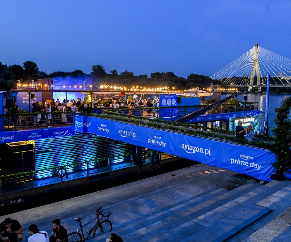 Event na barce Amazon Prime Day w Warszawie: tłum ludzi, oświetlenie, most w tle. Wieczorny plener z rowerem i grupą osób na nabrzeżu.