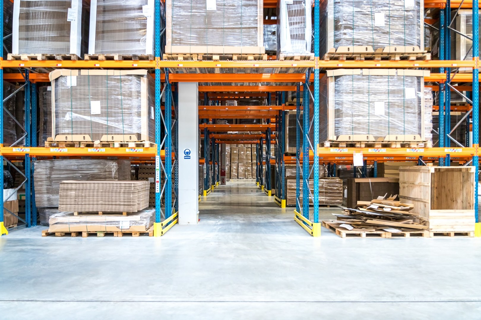 Wnętrze magazynu z regałami wypełnionymi paletami z towarem, widok w głąb korytarza między regałami, niebiesko-pomarańczowa konstrukcja regałów.
