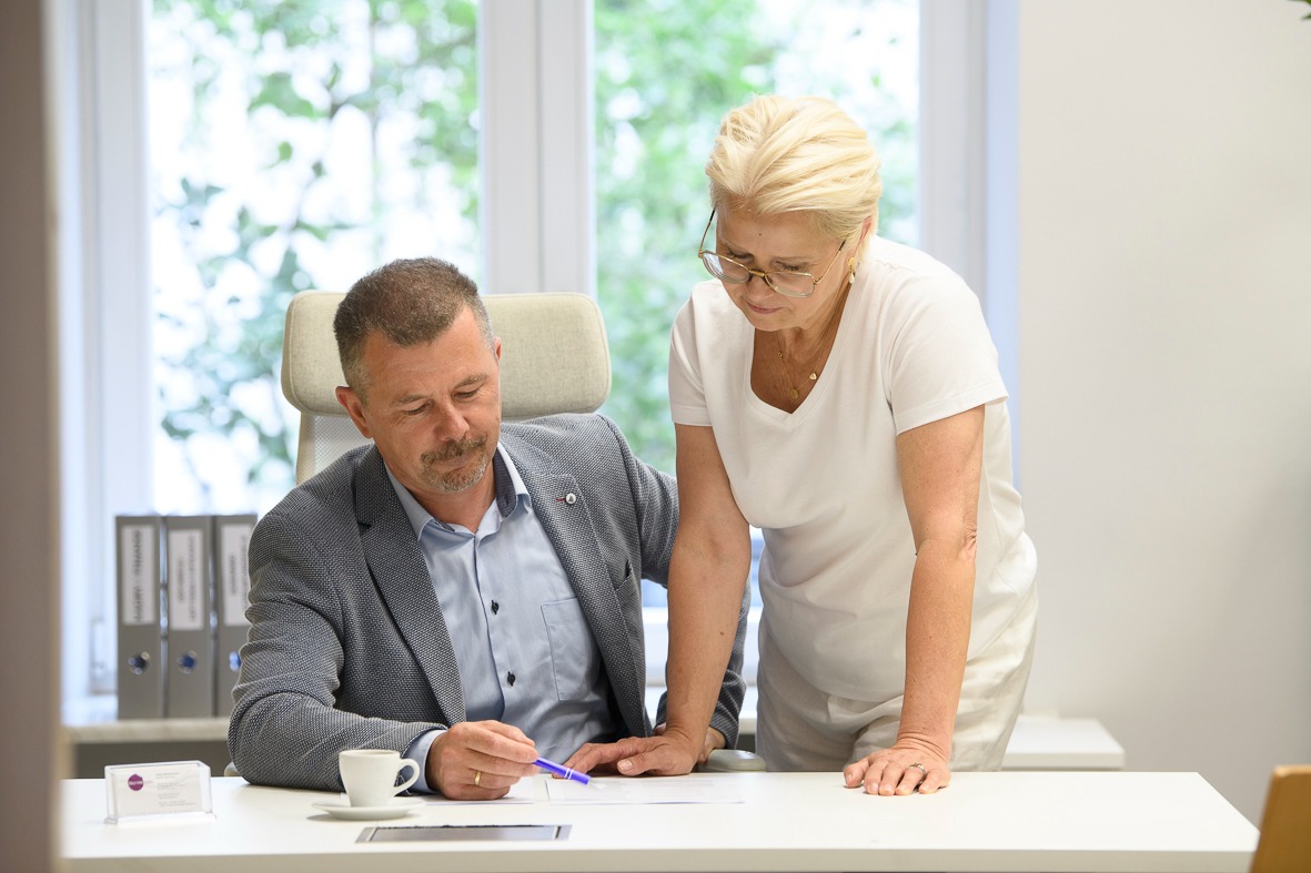 Dwie osoby pochylają się nad dokumentami na biurku w jasnym biurze. Mężczyzna w marynarce trzyma długopis, kobieta w okularach czyta. Widoczne segregatory i filiżanka kawy.