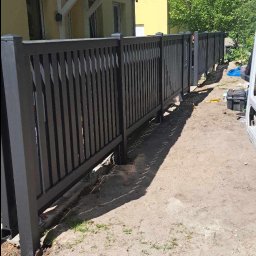 RB Błysk - Nowoczesne, grafitowe ogrodzenie panelowe z pionowymi szczebelkami, zamontowane przy żółtym domu z czerwonym dachem. Widoczny fragment białego samochodu dostawczego.