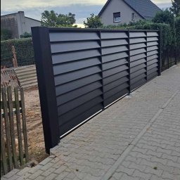 RB Błysk - Nowoczesne, poziome, czarne panele ogrodzeniowe z metalu, zamontowane na posesji. Widok z boku, z kostką brukową i fragmentem starego, drewnianego płotu.