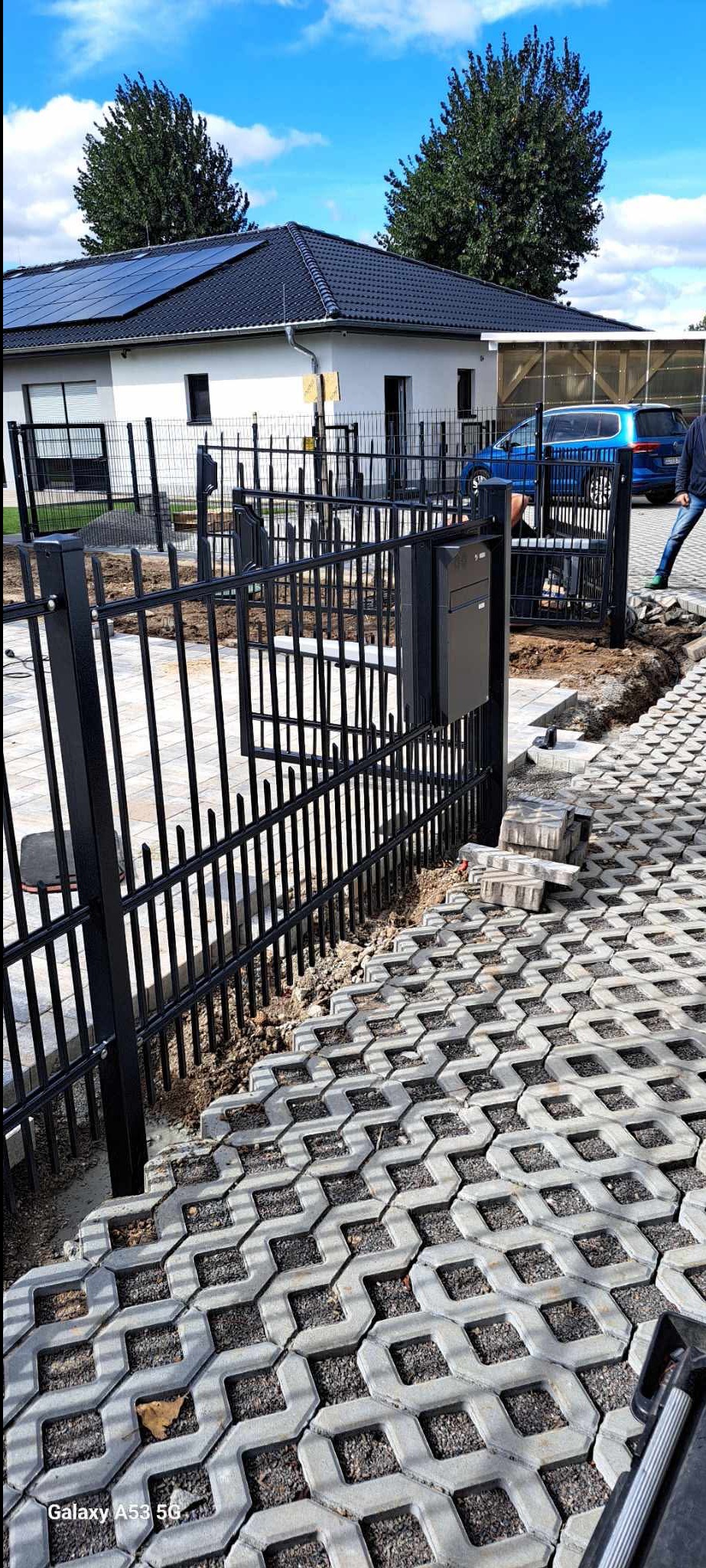Czarny metalowy płot z furtką i skrzynką na listy, w tle dom z panelami słonecznymi i fragment niebieskiego auta. Kostka ażurowa na podjeździe.