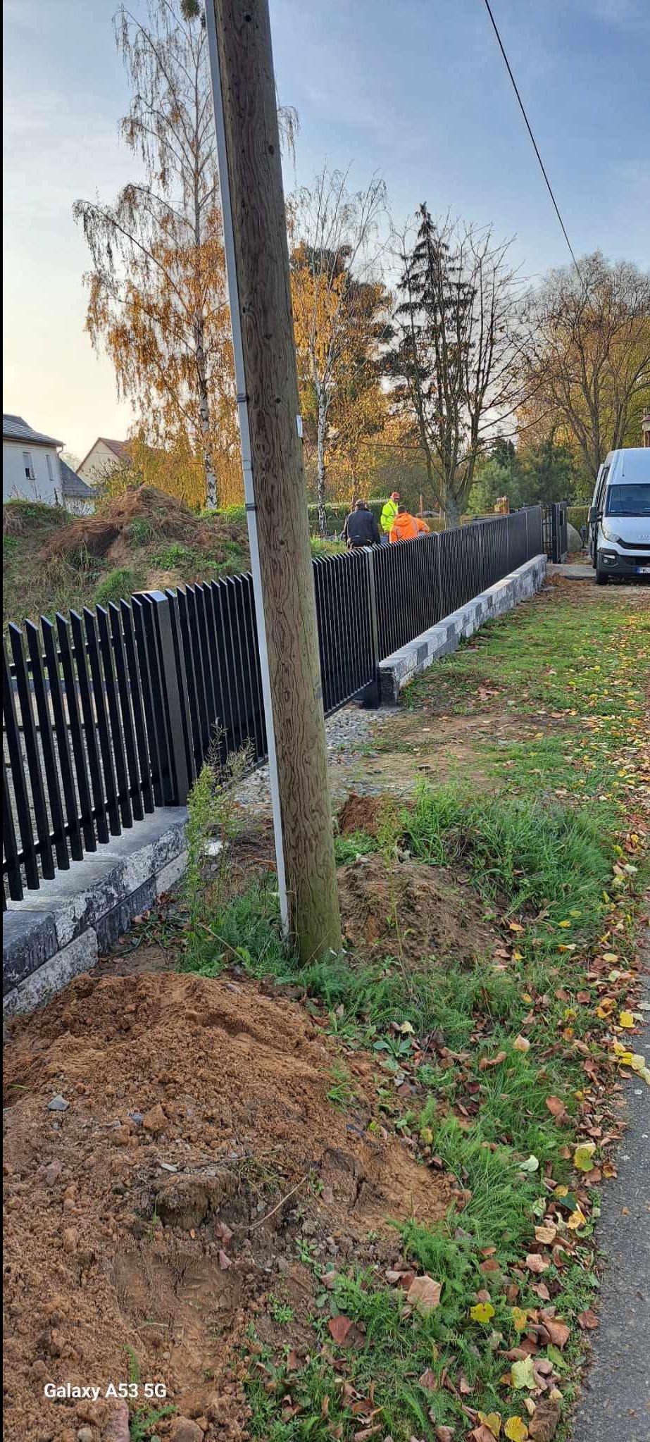 Nowoczesne, czarne ogrodzenie metalowe z pionowymi sztachetami, osadzone na betonowej podmurówce. W tle ekipa montażowa i samochód dostawczy. Widoczne ślady prac ziemnych.