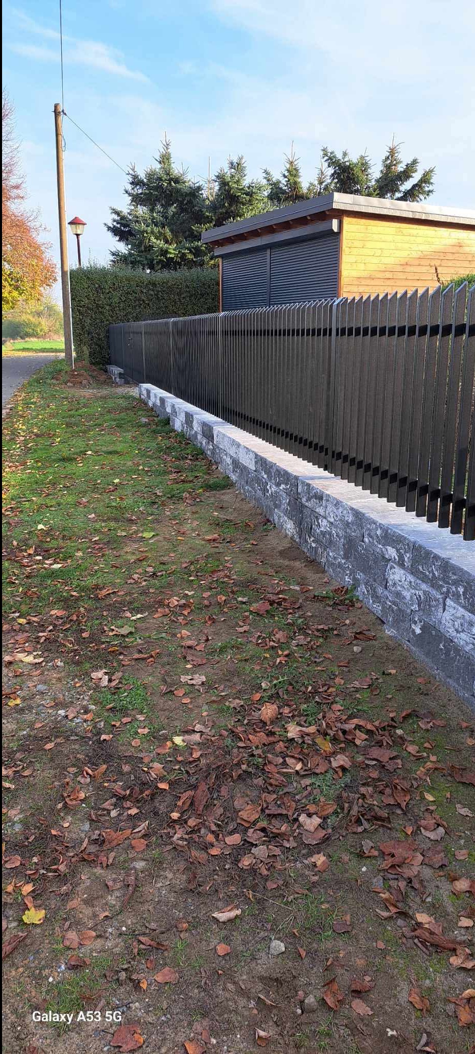 Nowoczesne ogrodzenie metalowe z pionowych sztachet, osadzone na niskim murku z kamienia łupanego. Jesienne liście na trawie, w tle budynek z szarą roletą.