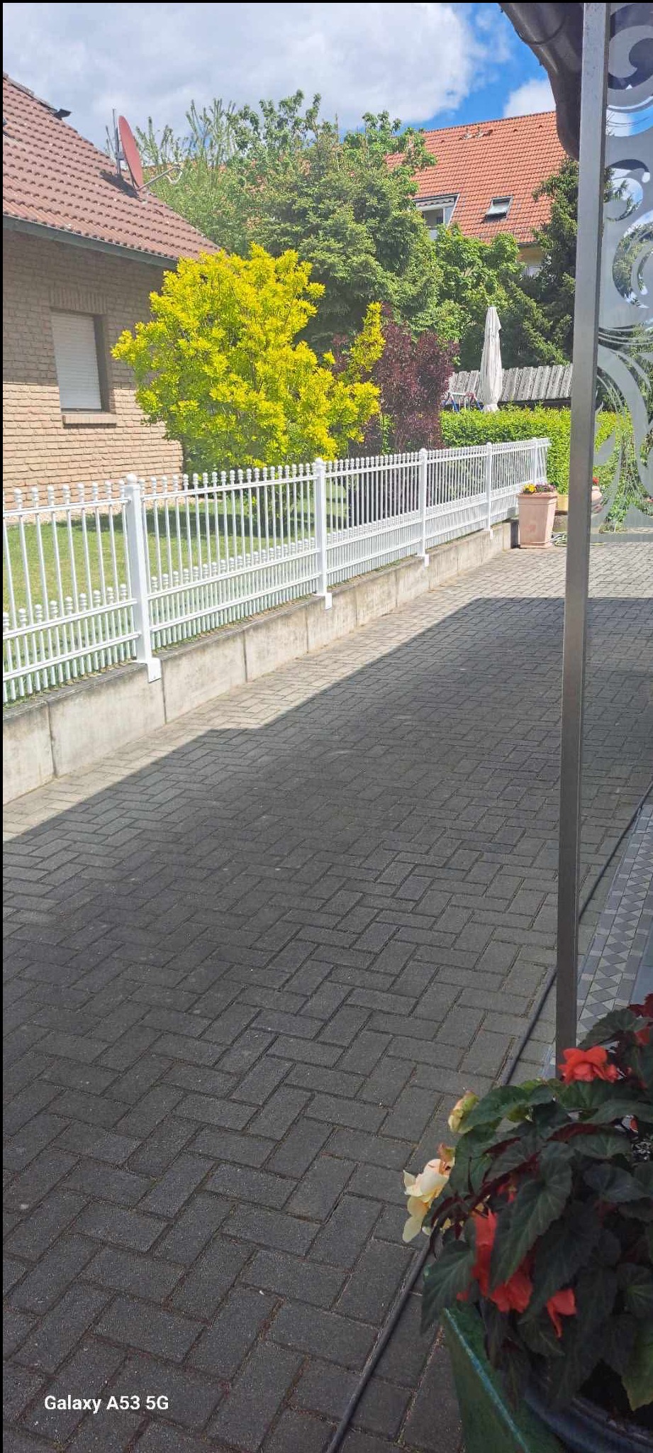 Białe, metalowe ogrodzenie panelowe z podmurówką, oddzielające posesję od chodnika. Widok na dom z czerwonym dachem i zielone drzewa w tle. Kostka brukowa na pierwszym planie.