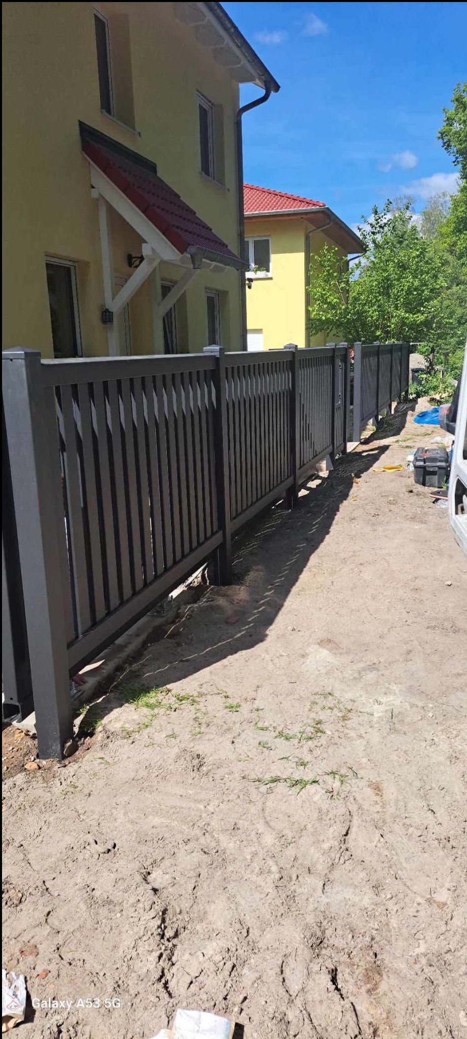 Nowoczesne, grafitowe ogrodzenie panelowe z pionowymi szczebelkami, zamontowane przy żółtym domu z czerwonym dachem. Widoczny fragment białego samochodu dostawczego.