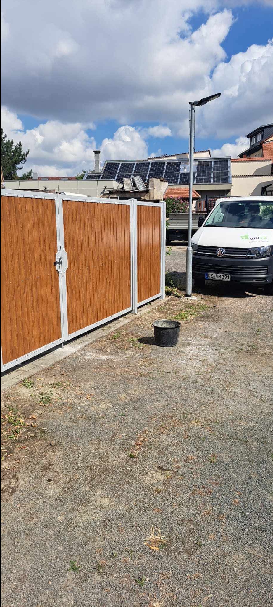 Nowoczesna brama z drewnopodobnymi panelami i białymi ramami, obok zaparkowany biały bus. W tle budynek z panelami słonecznymi na dachu.