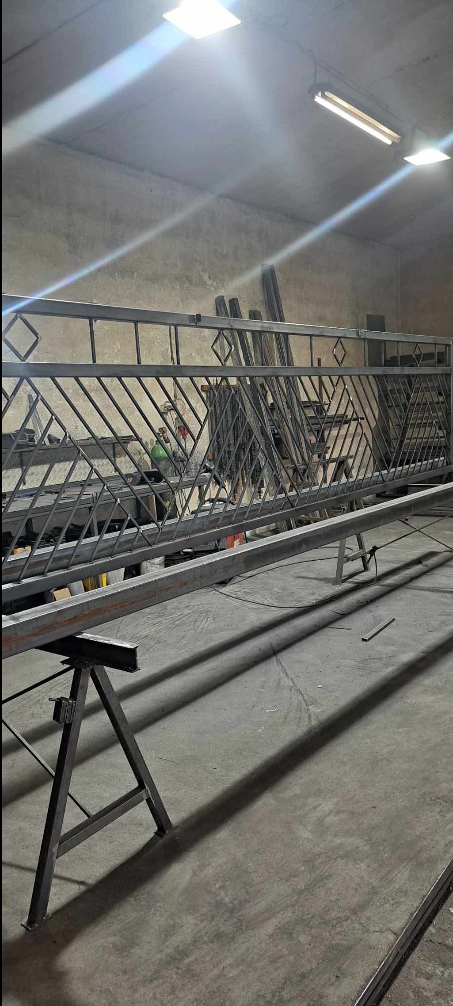 Surowy, metalowy fragment balustrady z geometrycznym wzorem w warsztacie, wsparty na kozłach. Widoczne narzędzia i materiały w tle, oświetlenie jarzeniowe.
