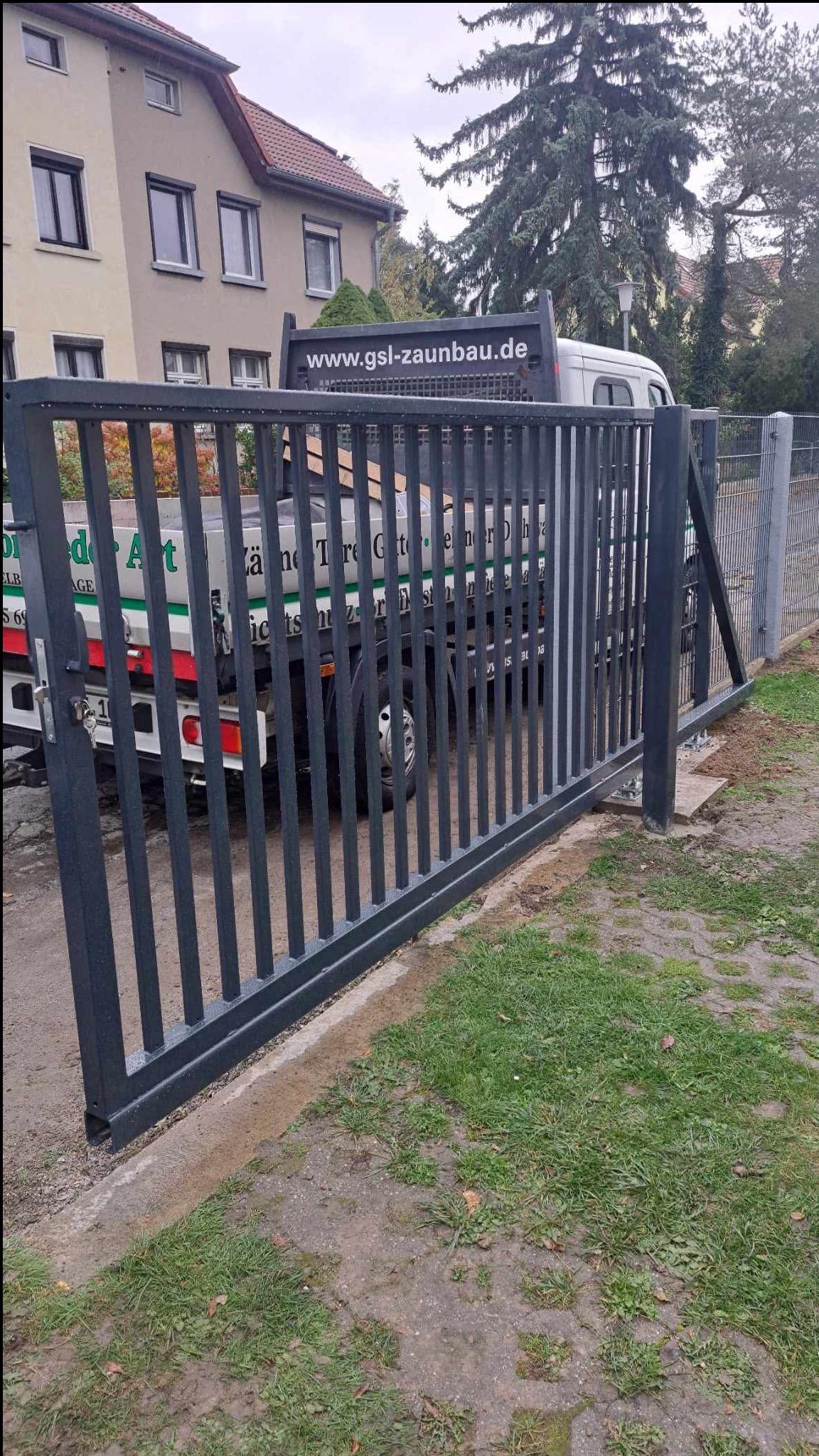 Nowoczesna, grafitowa brama przesuwna z pionowymi szczebelkami, zamontowana przy posesji. W tle widoczny samochód dostawczy z logo firmy.