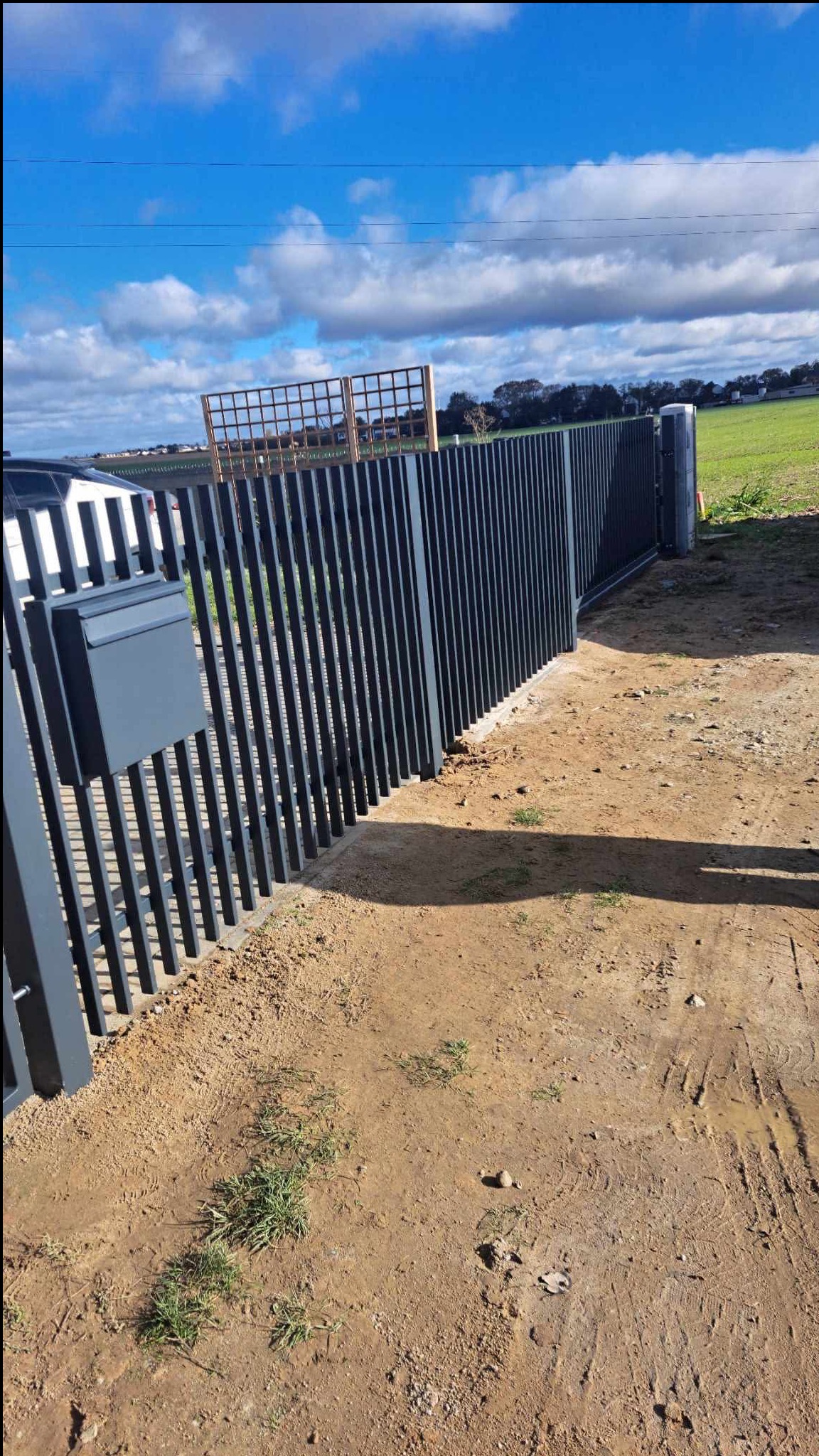 Nowoczesna, metalowa brama wjazdowa z pionowymi szczeblami i skrzynką na listy, na tle zielonego pola i błękitnego nieba z chmurami. Widoczna pergola w tle.