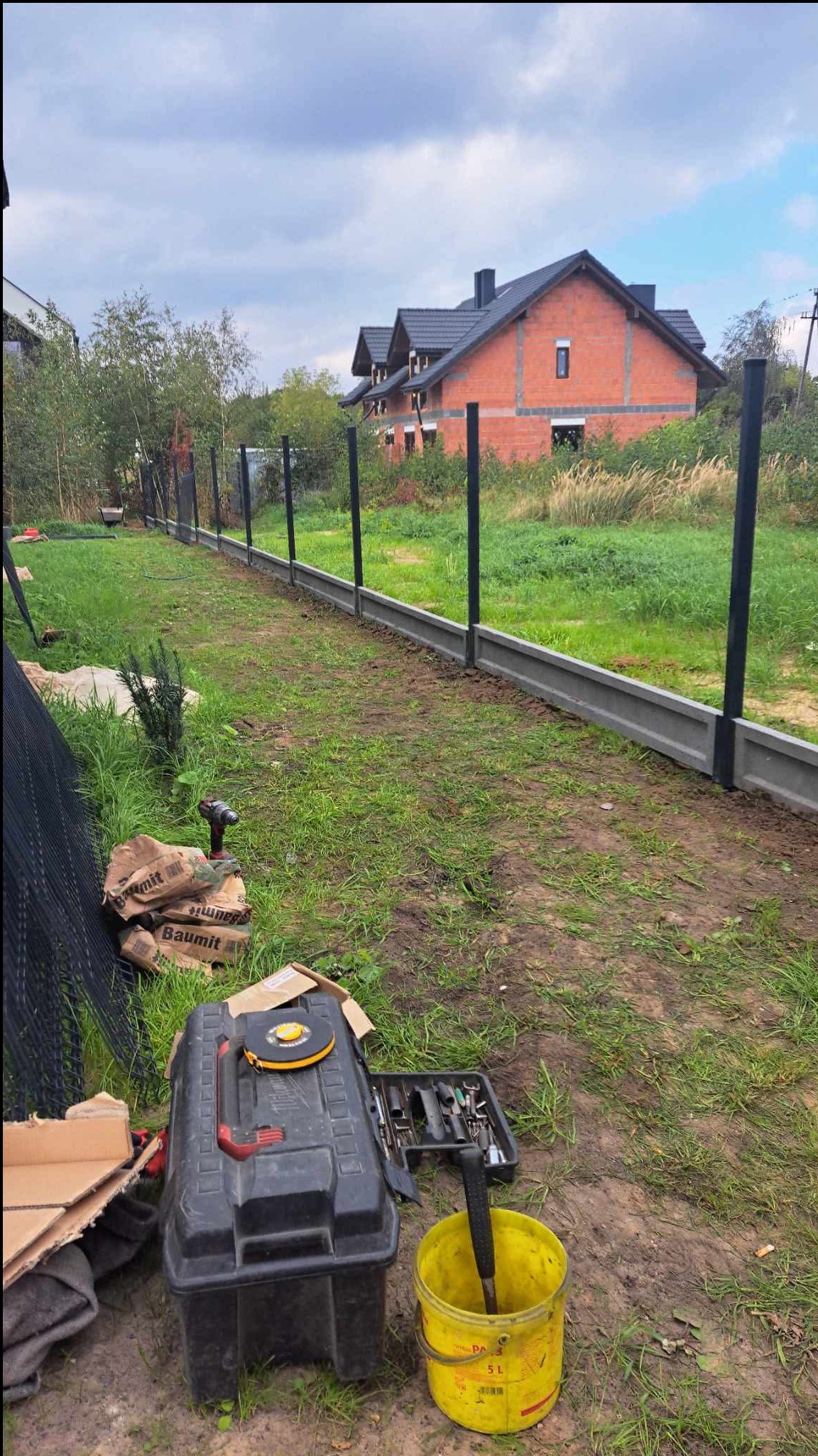 Montaż podmurówki i słupków ogrodzeniowych na działce z widokiem na dom. Widoczne narzędzia i materiały budowlane. Etap budowy ogrodzenia.